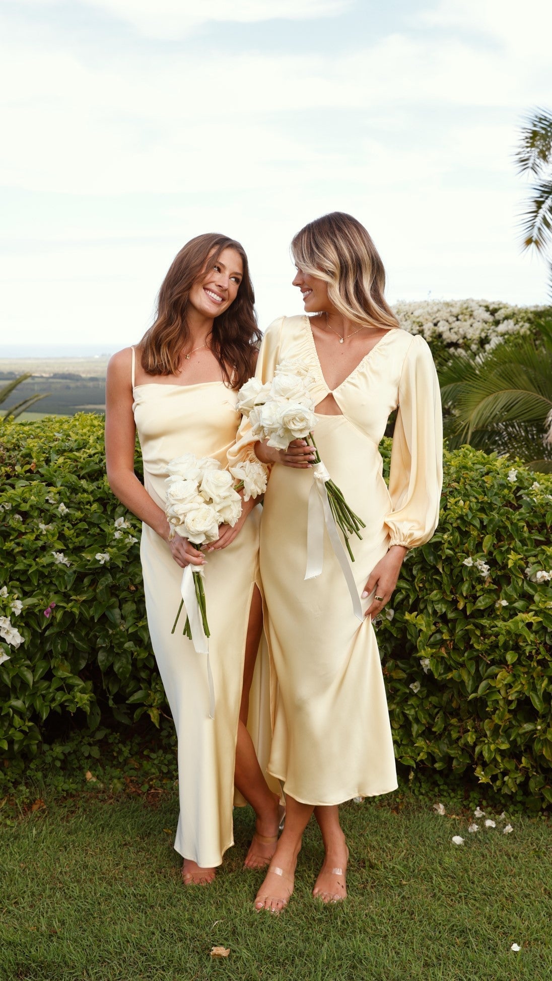 Two women, one in a yellow satin slip maxi dress and another in a long-sleeved yellow satin dress, holding bouquets.