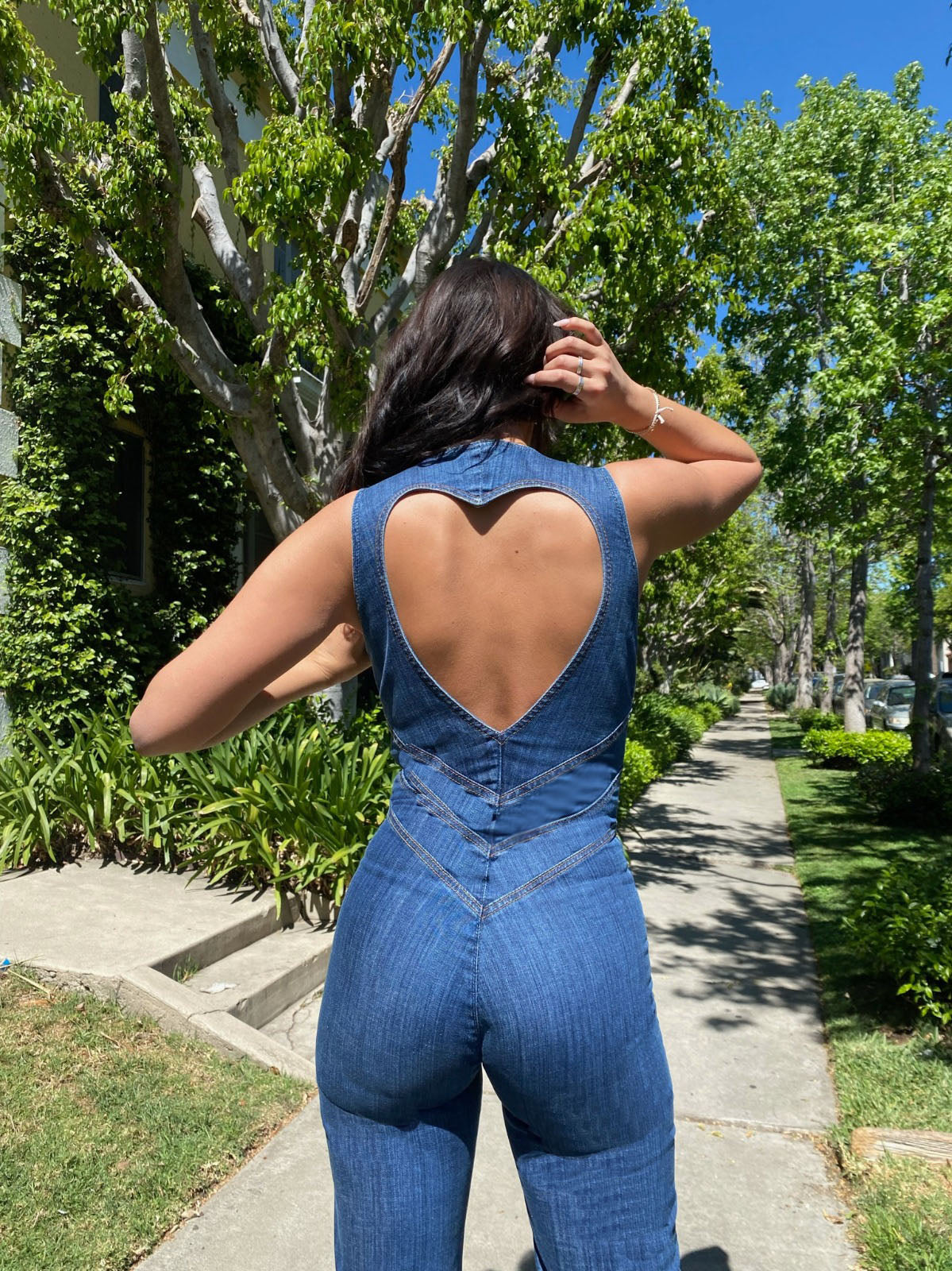 Back view of a woman in a dark wash denim jumpsuit, showcasing the heart-shaped cutout and V-stitch detailing, on a tree-lined street.