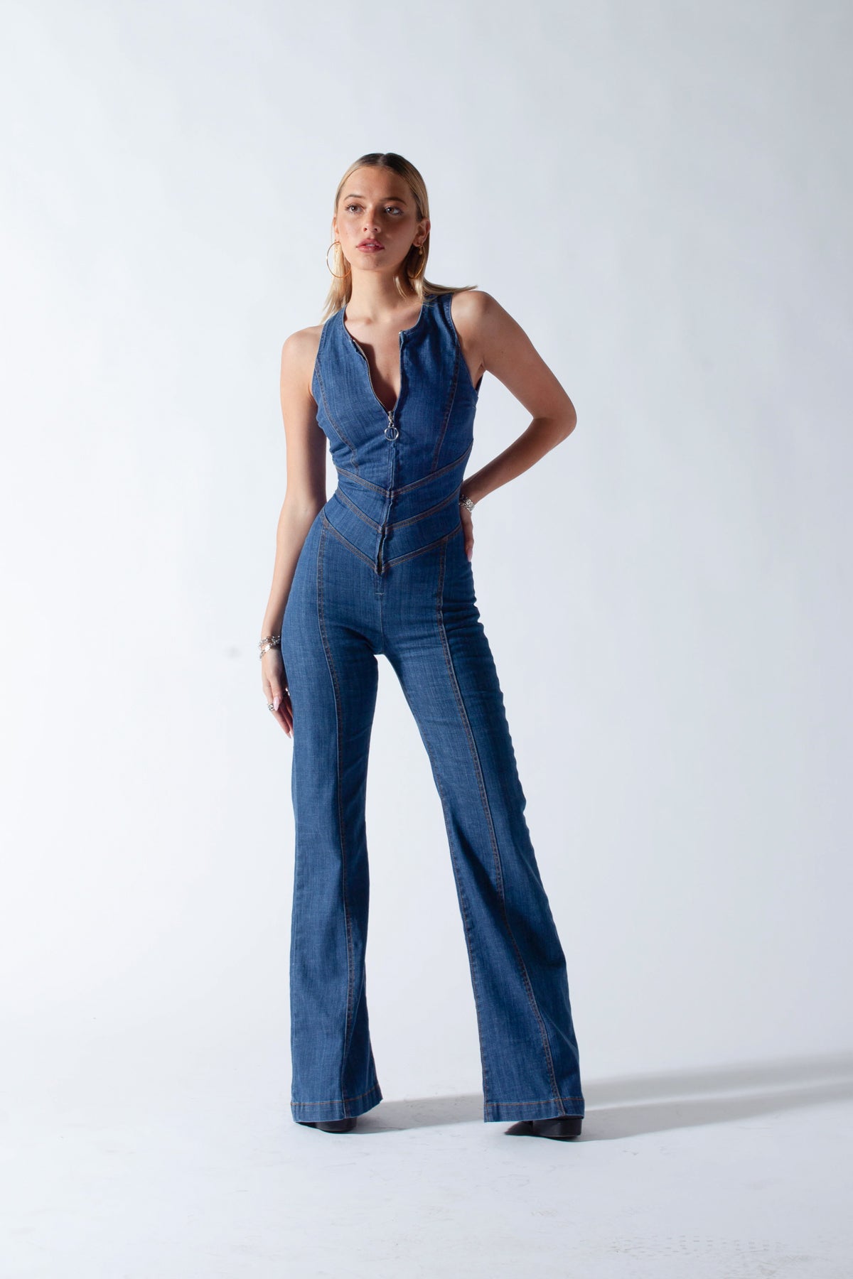 Studio shot of a woman posing in a form-fitting dark wash denim jumpsuit with a front zipper and flared bell-bottom legs.