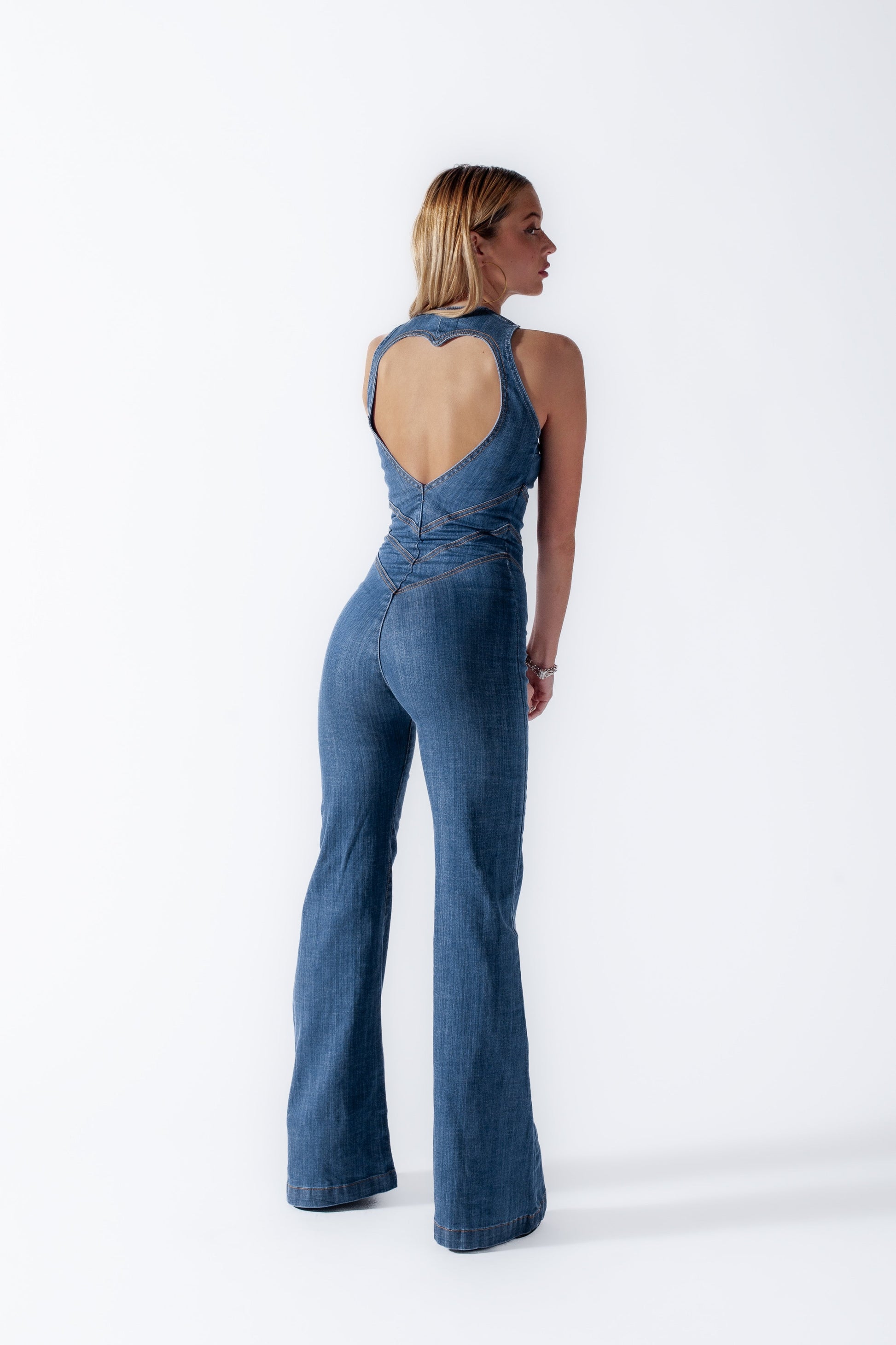 Rear view of a woman in a dark wash denim jumpsuit, showing the heart-shaped back cutout and seam detailing on the flared pants.