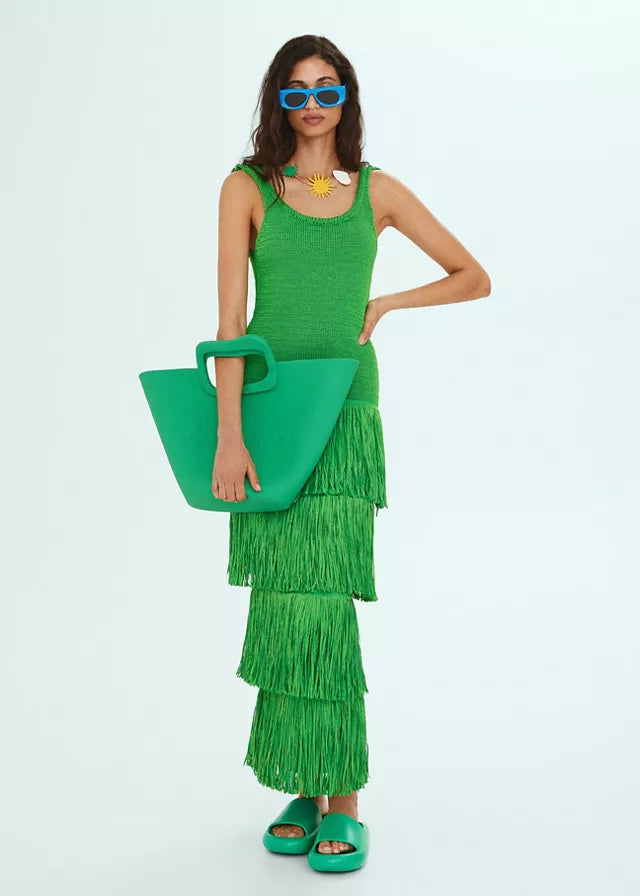 Front full shot of a model wearing a green tiered fringe midi dress, blue sunglasses, and holding a green tote bag.