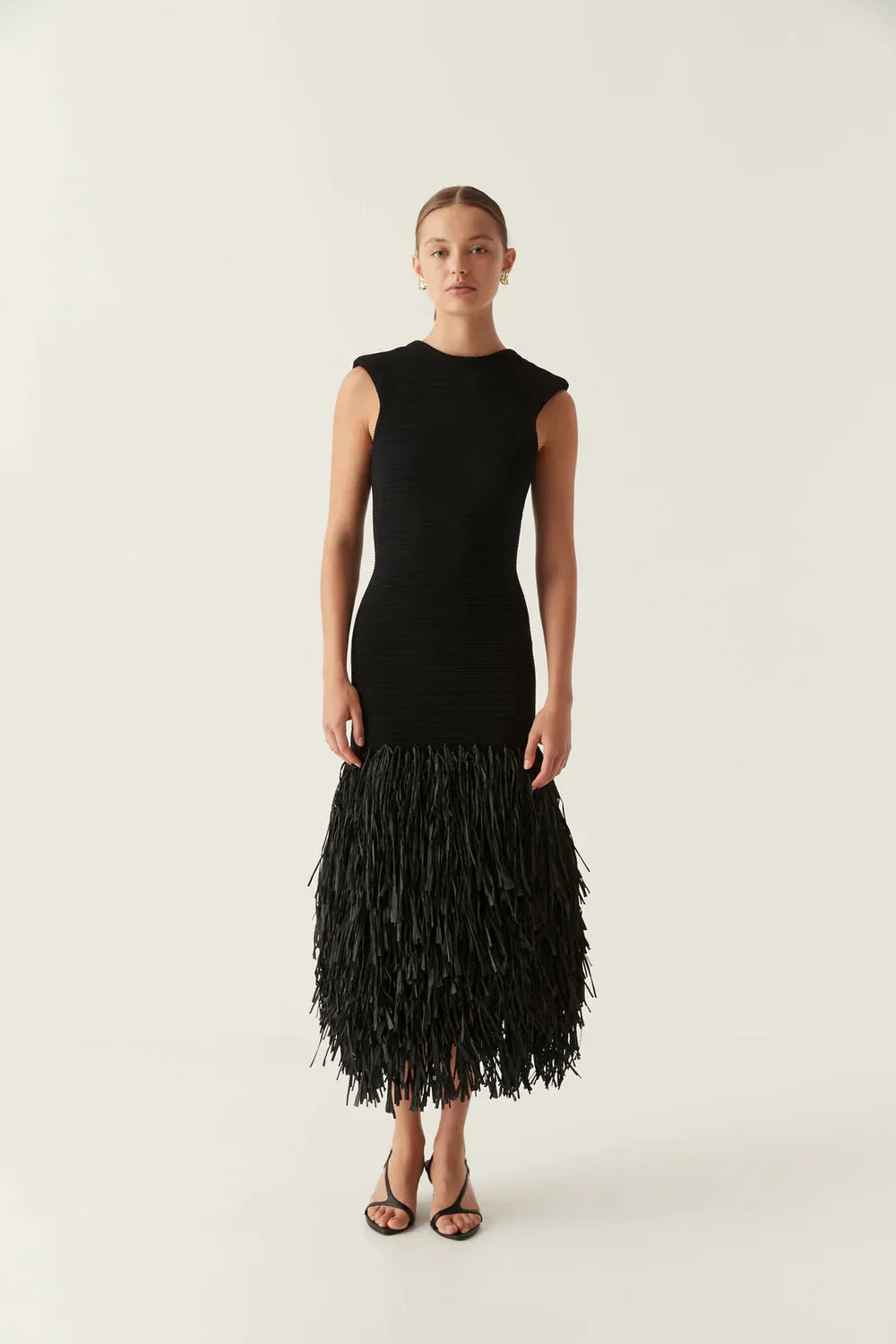 Black sleeveless midi dress with ribbed bodice and long fringe details on the skirt, worn with black heels.