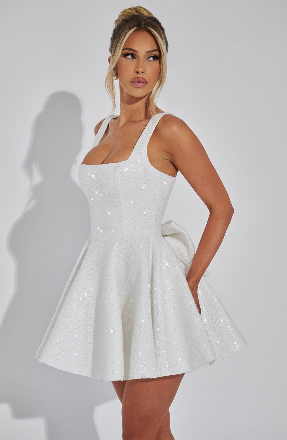 Side profile of a white sequin mini dress, showcasing the square neckline, flared skirt, and subtle bow detail at the hip.