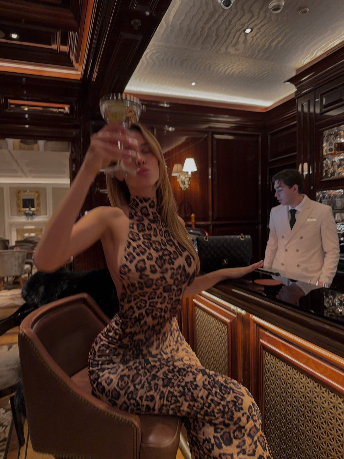 Woman in a leopard print maxi dress with a halter neckline, holding a cocktail glass at a bar.
