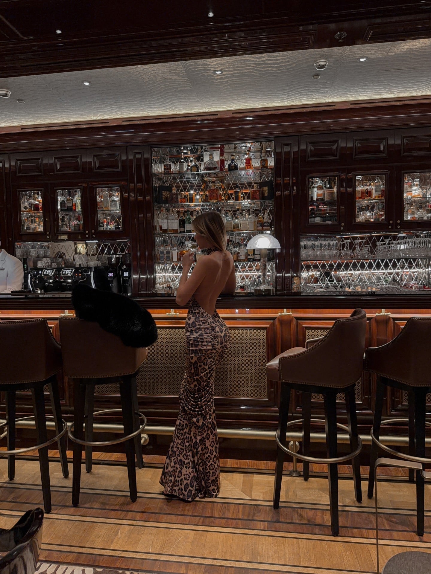 Back view of a woman in a long leopard print maxi dress with ruched details, standing at a bar.