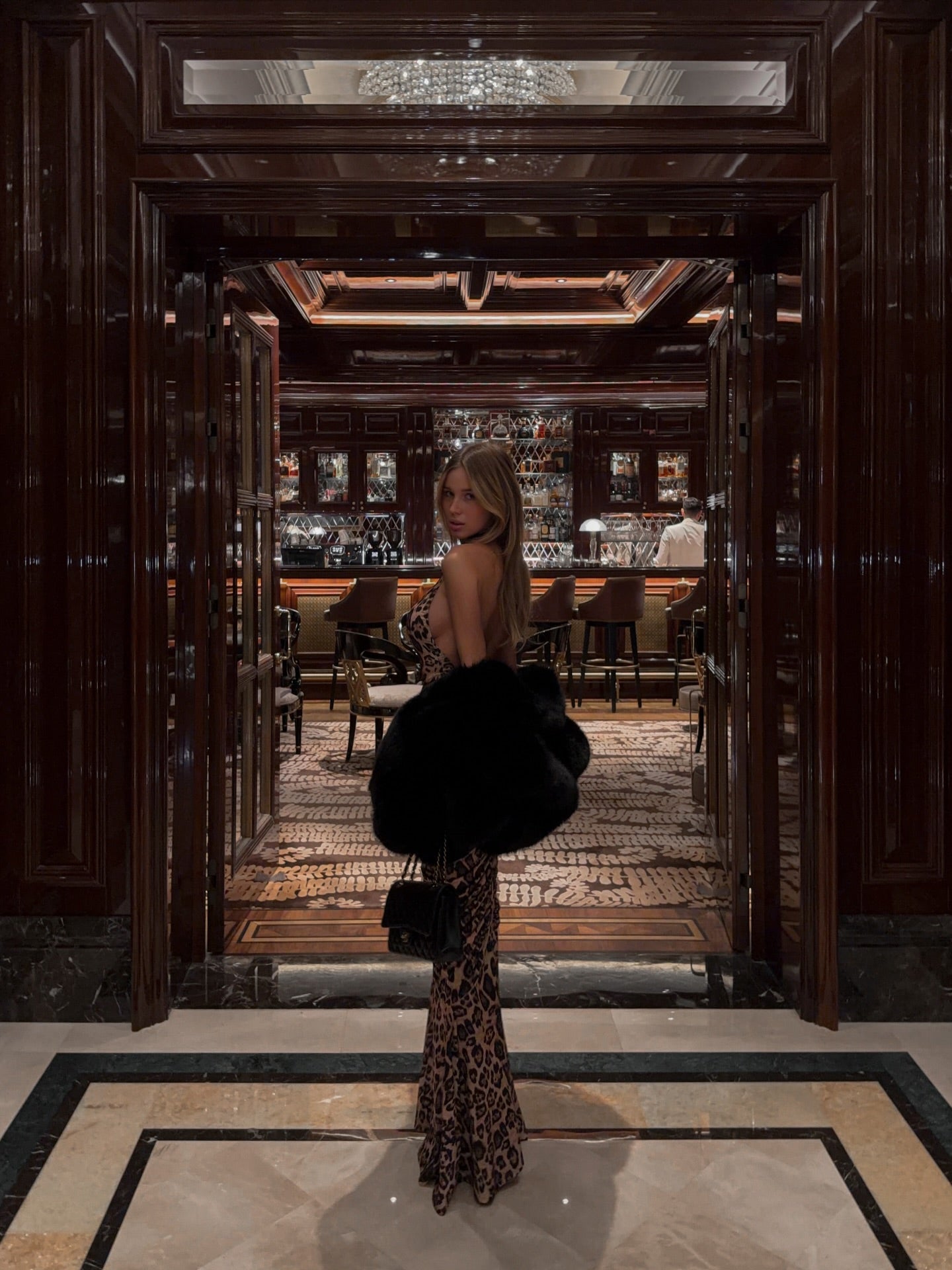 Woman in a leopard print maxi dress and black faux fur coat, looking back from a doorway.
