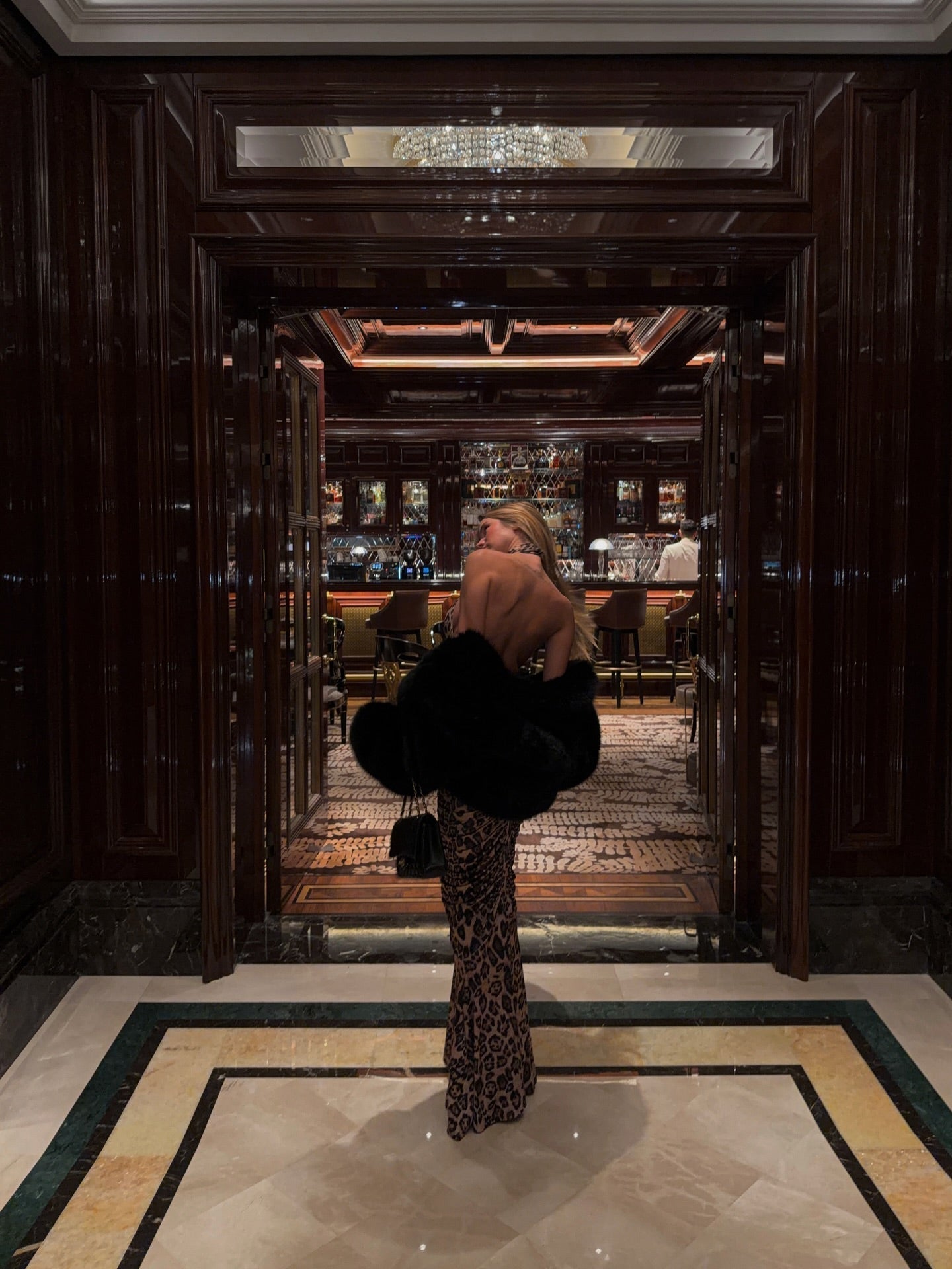 Rear view of a woman in a leopard print maxi dress, with a black faux fur coat draped over her arms.
