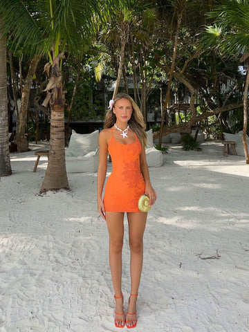 Front view of an orange sequin mini dress with spaghetti straps and floral embellishments, worn on a sandy beach.