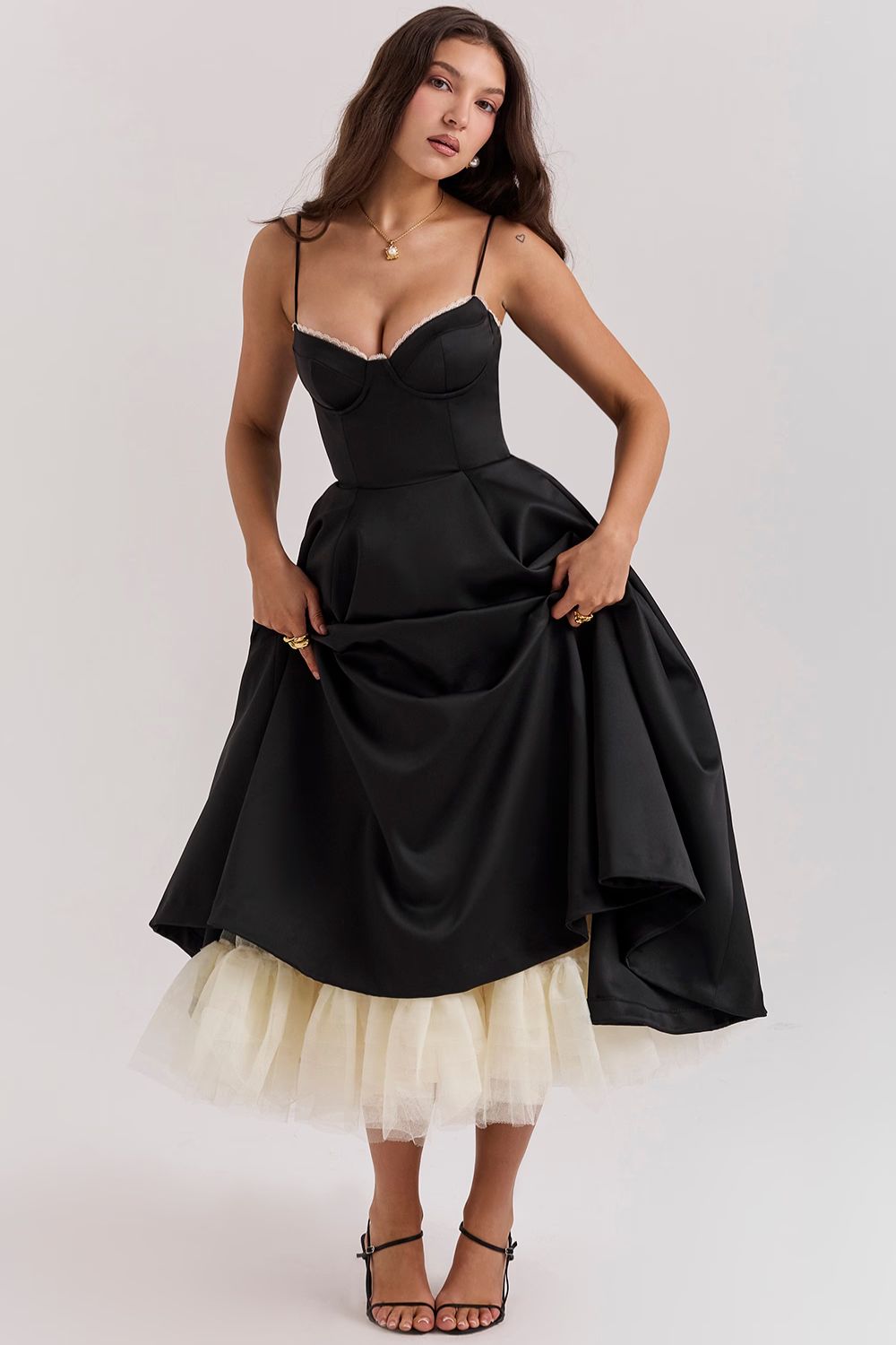 Full body view of a woman wearing a black corset midi dress with a full skirt and visible cream tulle underskirt, holding up the skirt slightly.