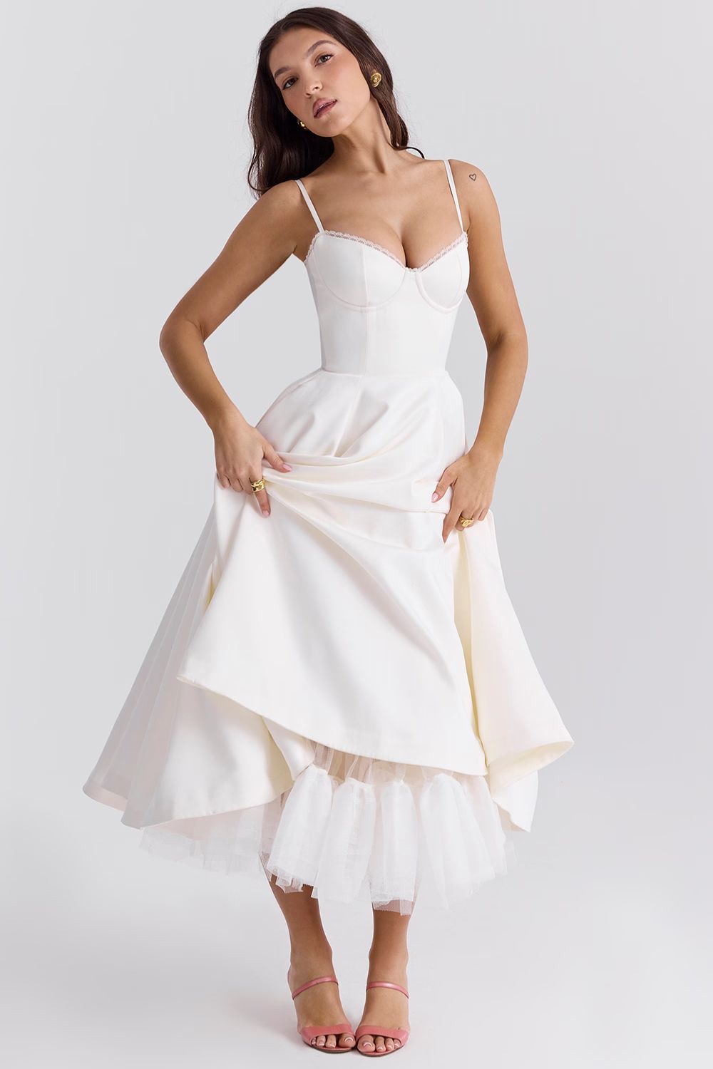 Full body shot of a woman in a white Angela corset midi dress, lifting the skirt to reveal a voluminous white tulle underskirt.