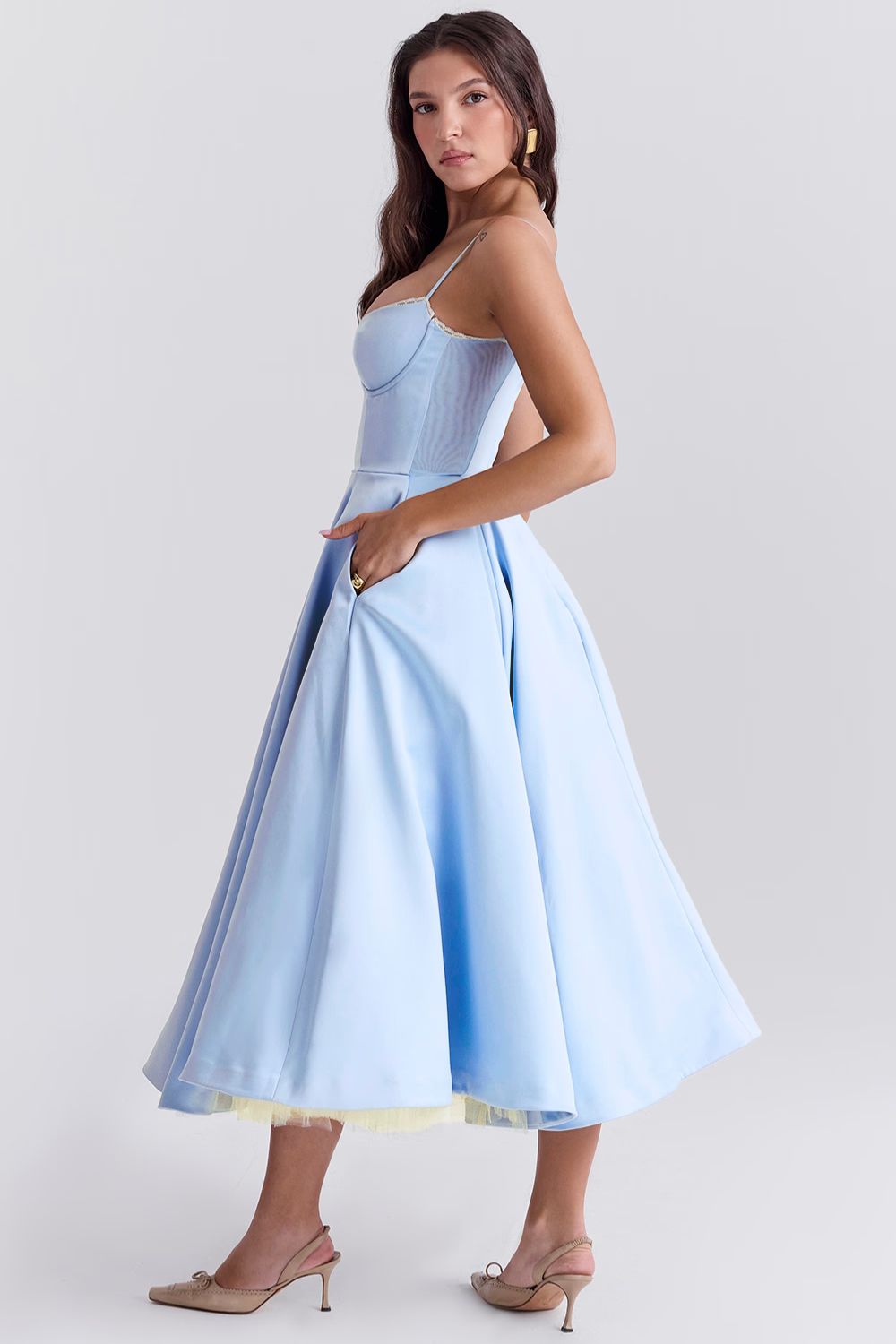 Side profile of a woman in a light blue Angela corset midi dress, with her hand in the dress pocket, revealing a yellow tulle underskirt.