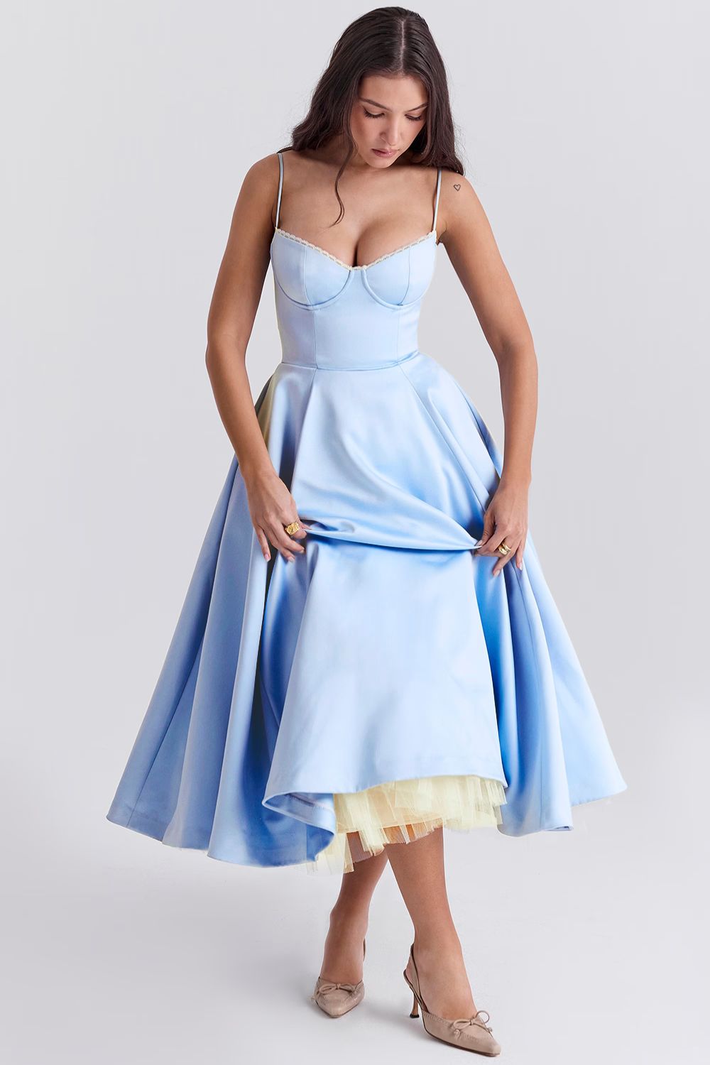 Front view of a woman wearing a light blue Angela corset midi dress, holding up the skirt to display the yellow tulle underskirt.