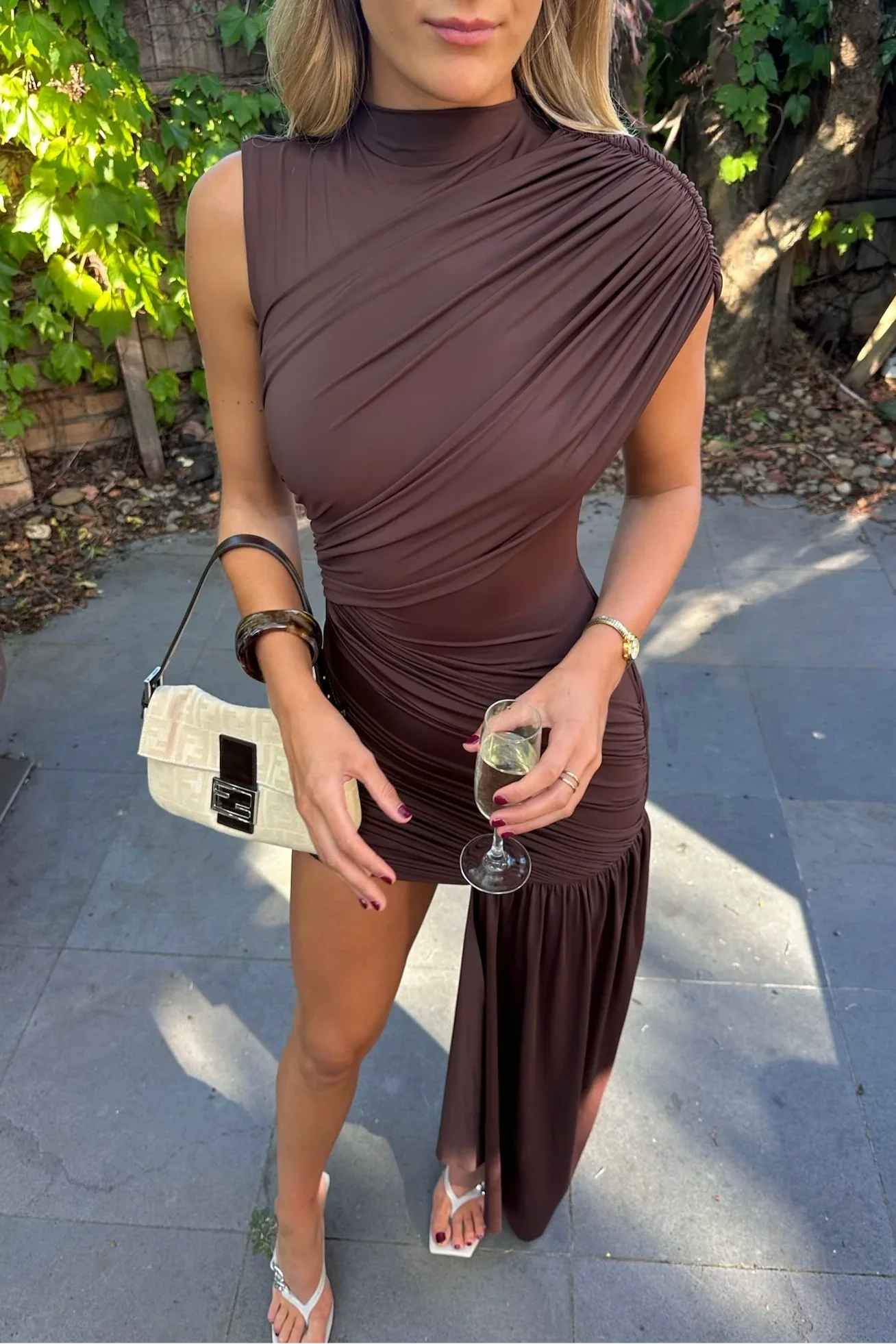 Brown Aria Ruched Mini Dress with high neckline, sleeveless design, and asymmetrical draped skirt, paired with white heels.