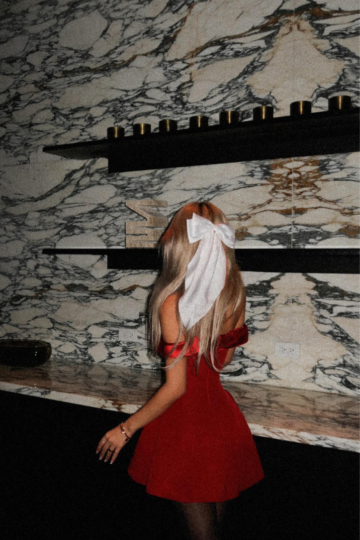 Back view of a blonde woman wearing a red off-shoulder mini dress, with a stylish white bow in her hair.