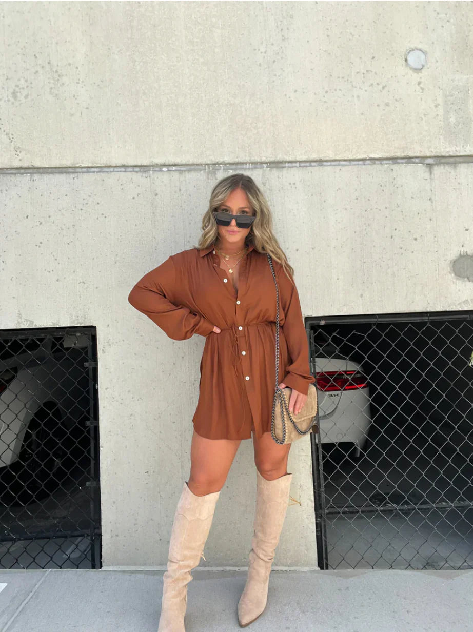 Full length view of the Belle Button Mini Romper in a rich rust brown color, paired with tall cream-colored boots and sunglasses.