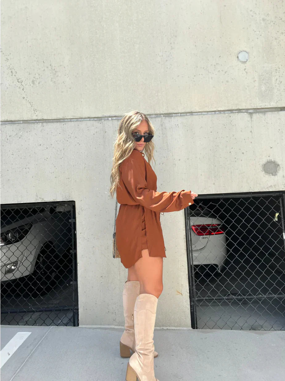Side profile of the rust brown Belle Button Mini Romper, showing the loose-fitting top and cinched waist, with tall cream boots.
