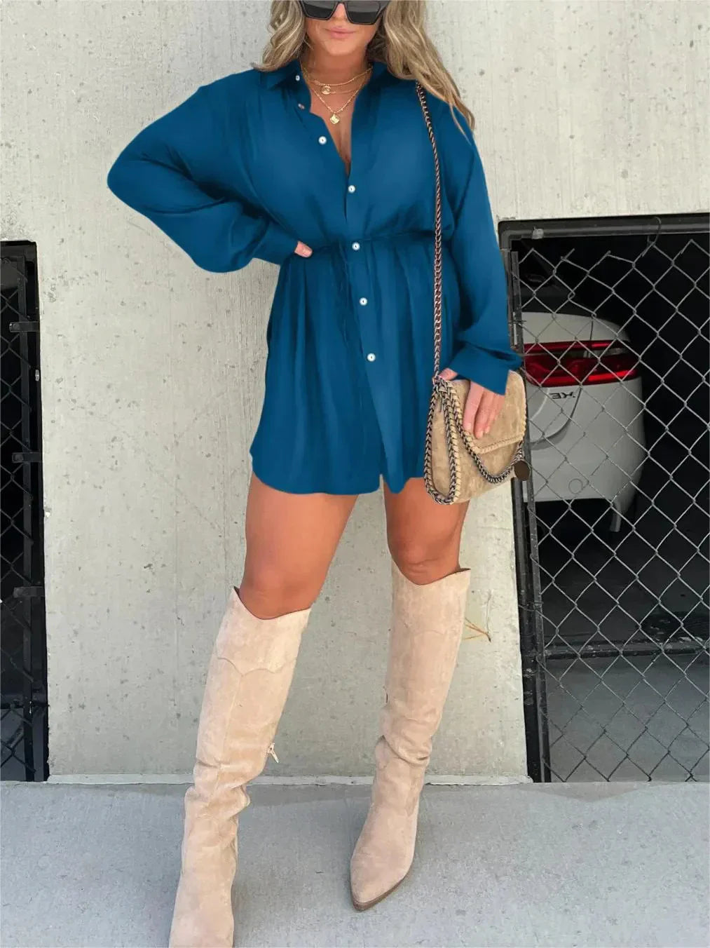 Teal blue Belle Button Mini Romper featuring a collared design and cinched waist, styled with tall beige boots and a chain-strap bag.