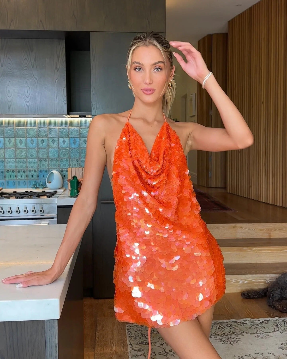 Orange sequin cowl halter mini dress on a woman leaning on a kitchen counter, looking away for women.
