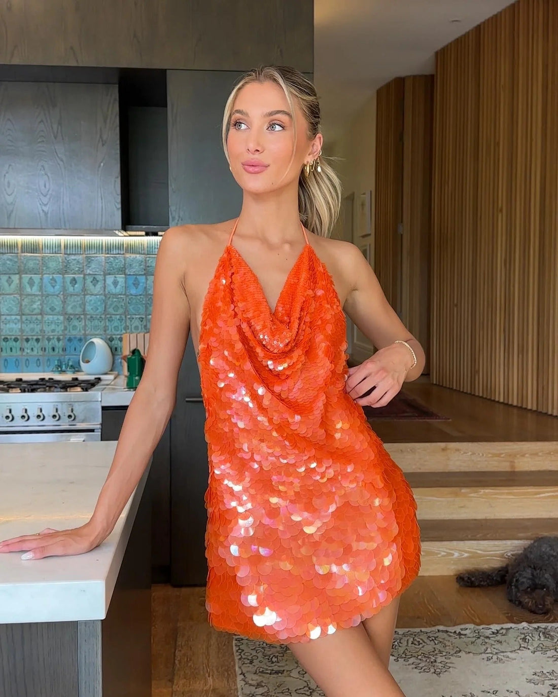 Orange sequin cowl halter mini dress on a woman leaning on a kitchen counter, looking away for women.