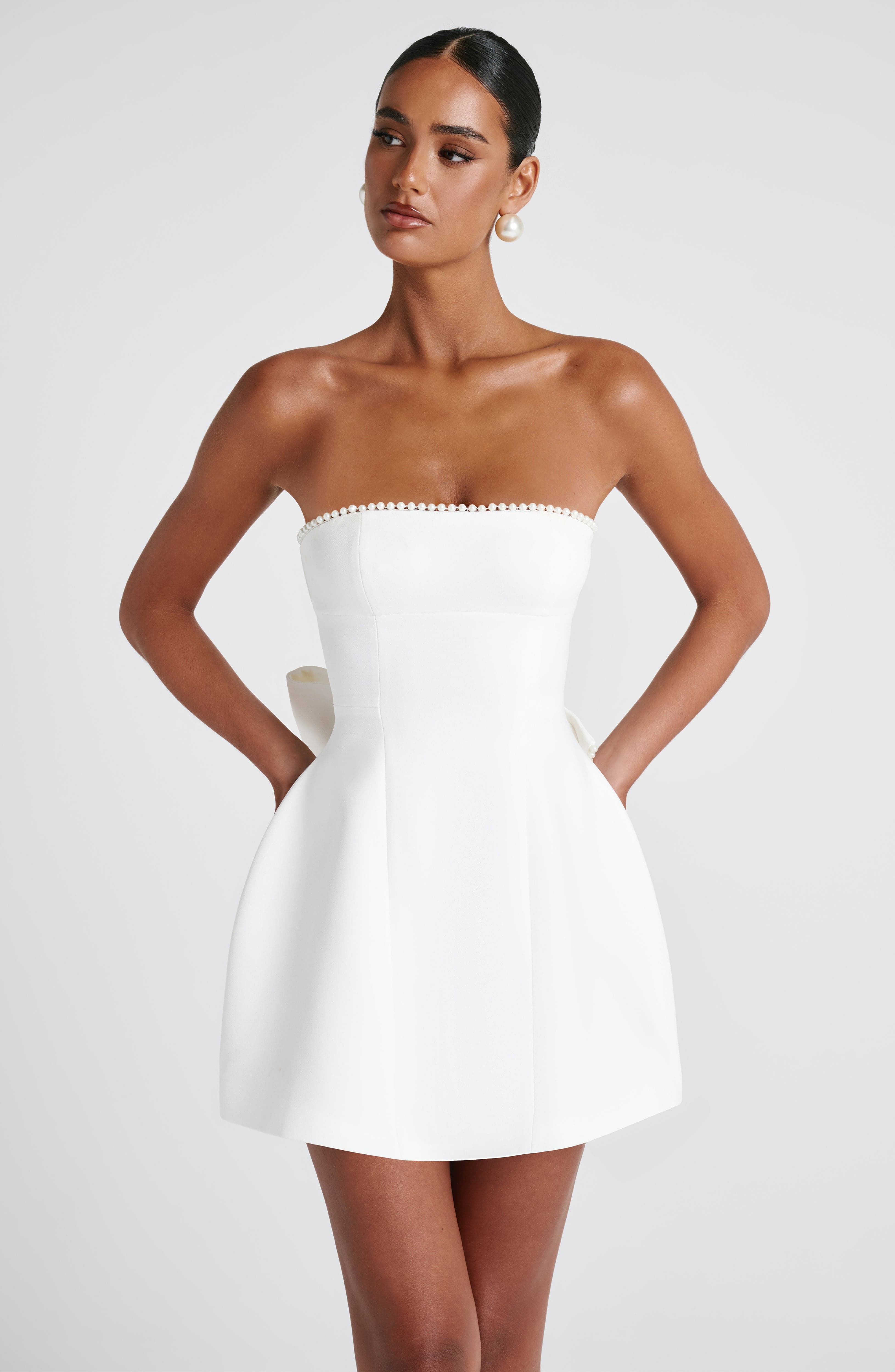 Front view of the white Calliope Pearl Bow Mini Dress, hands in pockets, highlighting the pearl neckline and structured fit.