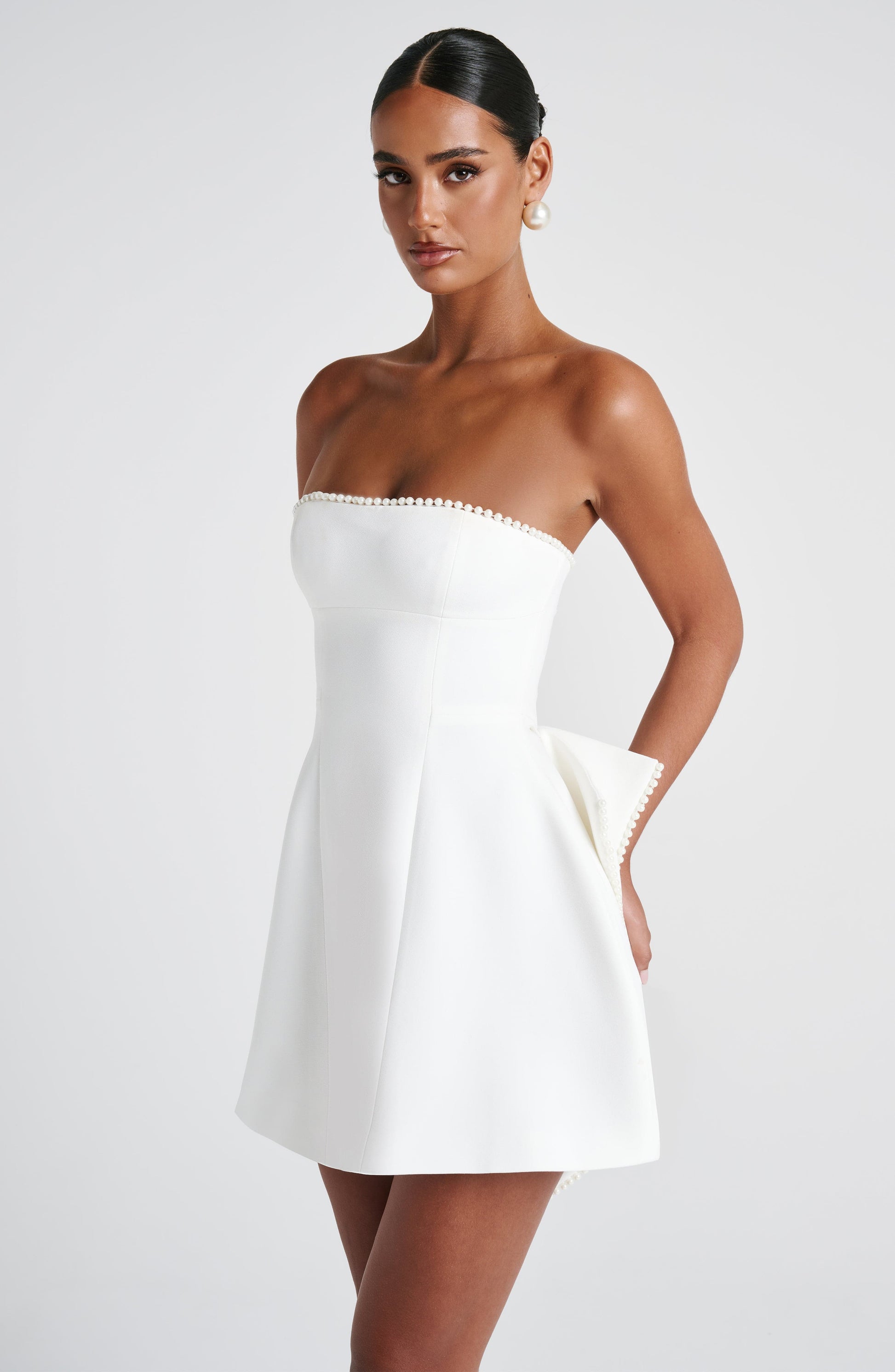 Side profile of the white Calliope Pearl Bow Mini Dress, hands in pockets, revealing a subtle side bow detail.