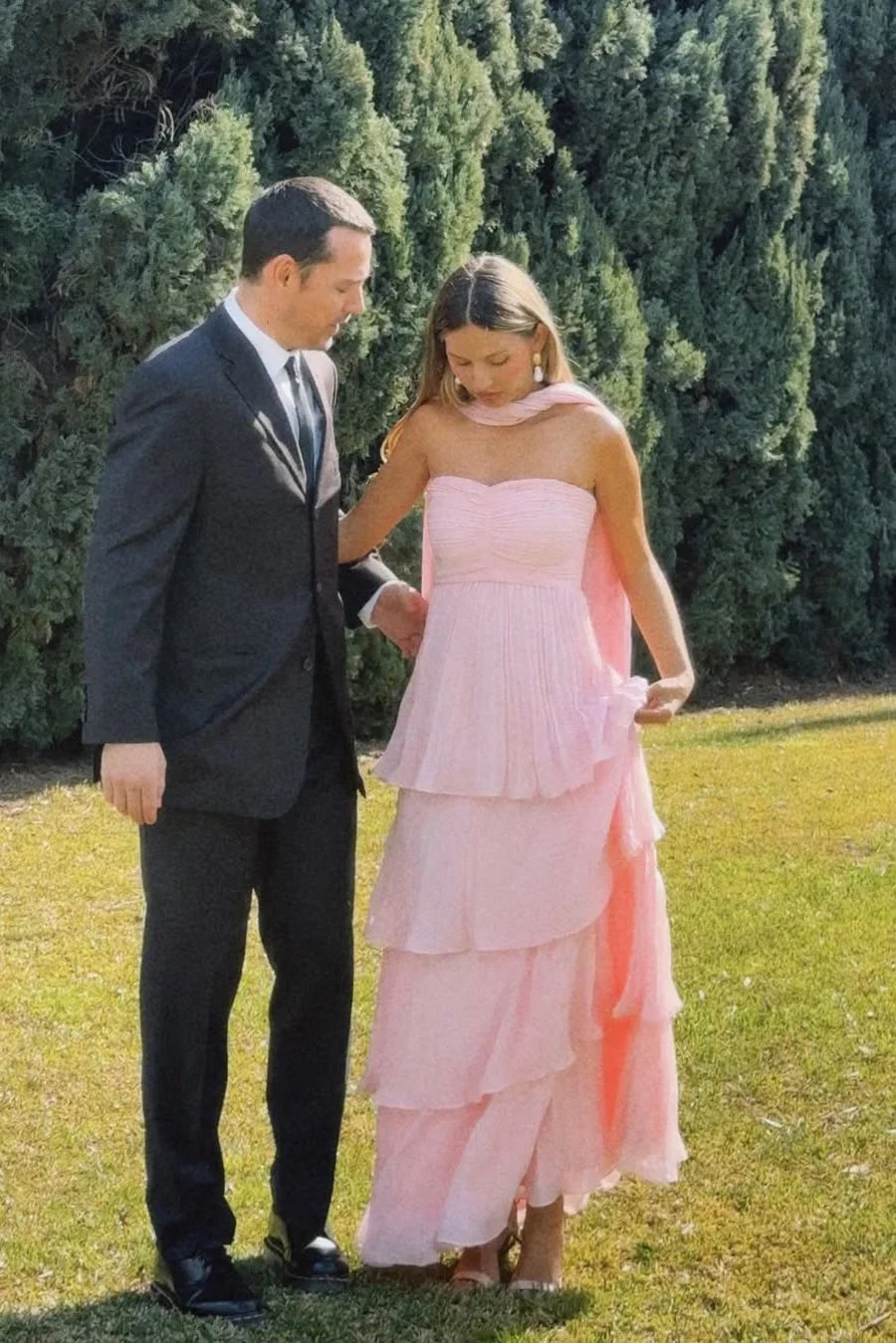 Full length view of a light pink tiered maxi dress with a strapless top and pleated skirt, worn with a man in a dark suit