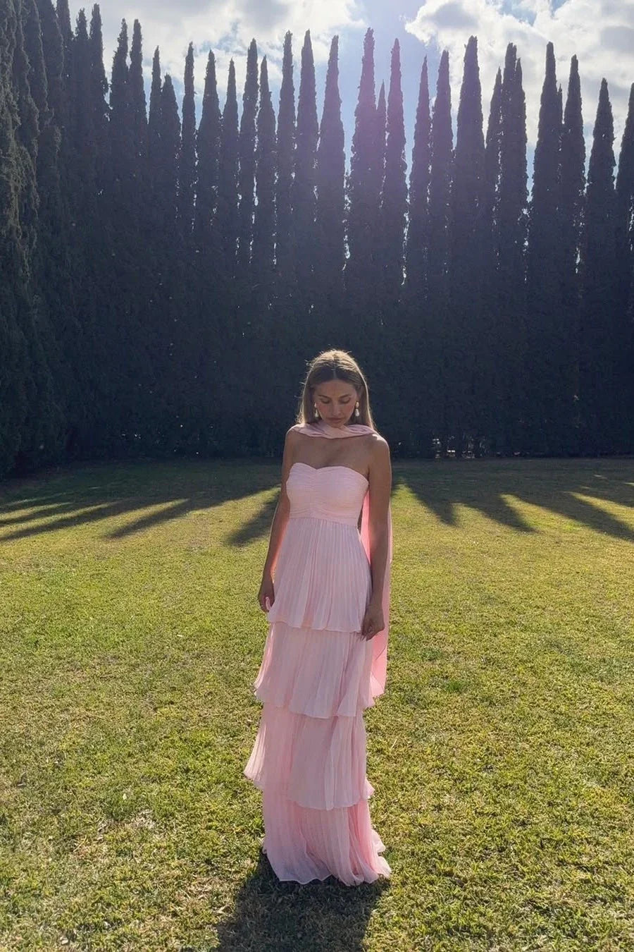 Full length view of a pink tiered maxi dress with a strapless, ruched bodice and a flowing pleated skirt in a garden