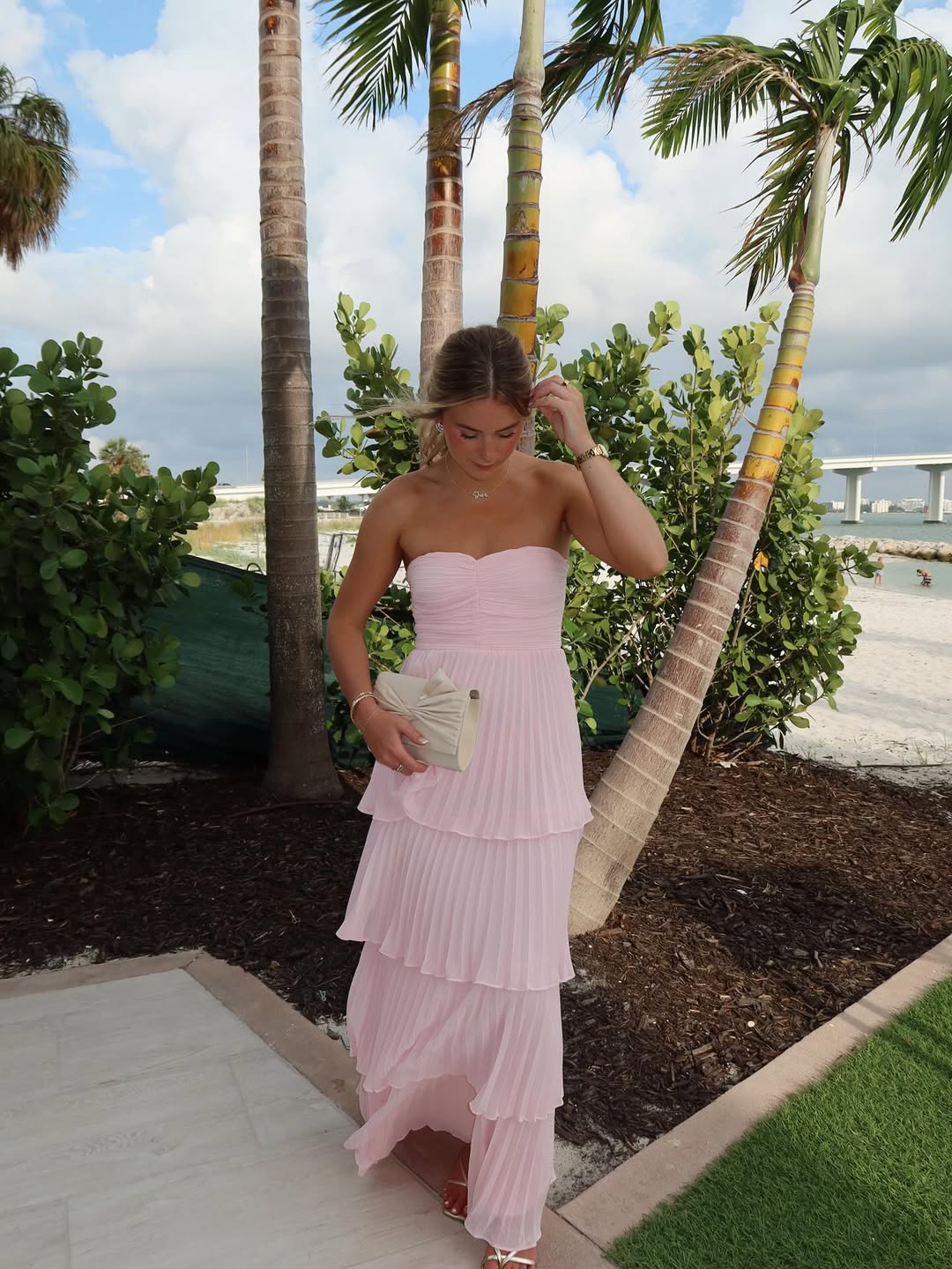 Outdoor shot of a woman in a light pink pleated tiered maxi dress, featuring a strapless design and a white clutch