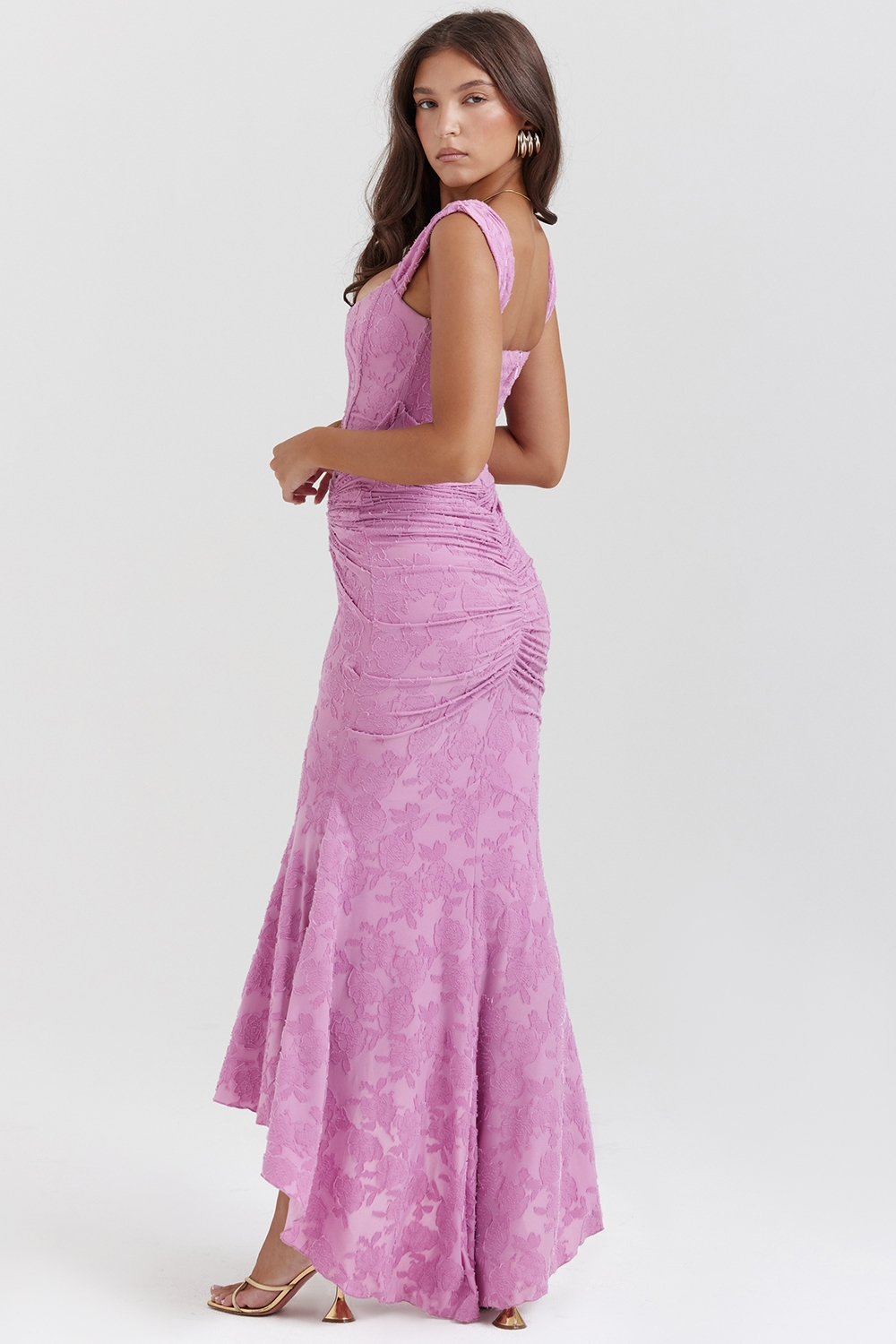 Side view of an orchid purple floral jacquard corset maxi dress with a ruched skirt and a high-low hem, styled with gold heeled sandals.