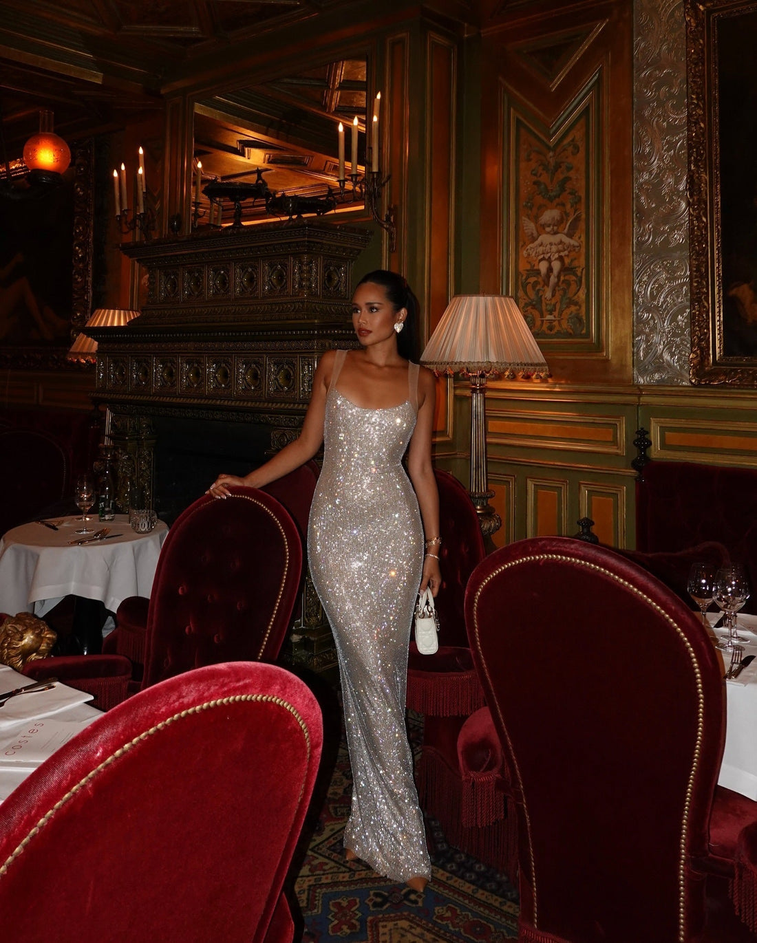 Shimmering Caroline Sequin Maxi Dress in silver with delicate straps, worn by a woman holding a wine glass on a red velvet couch.