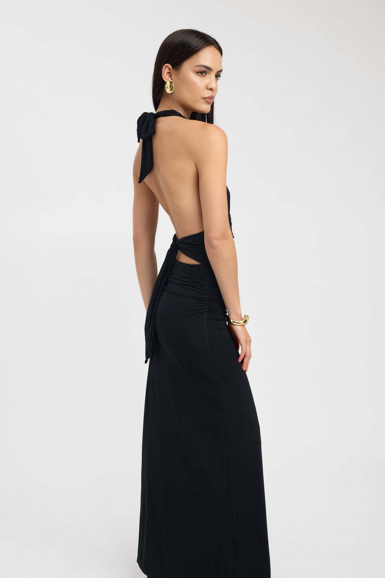 Side profile of a black ruched maxi dress, highlighting the open back, tie detail, and gathered fabric.