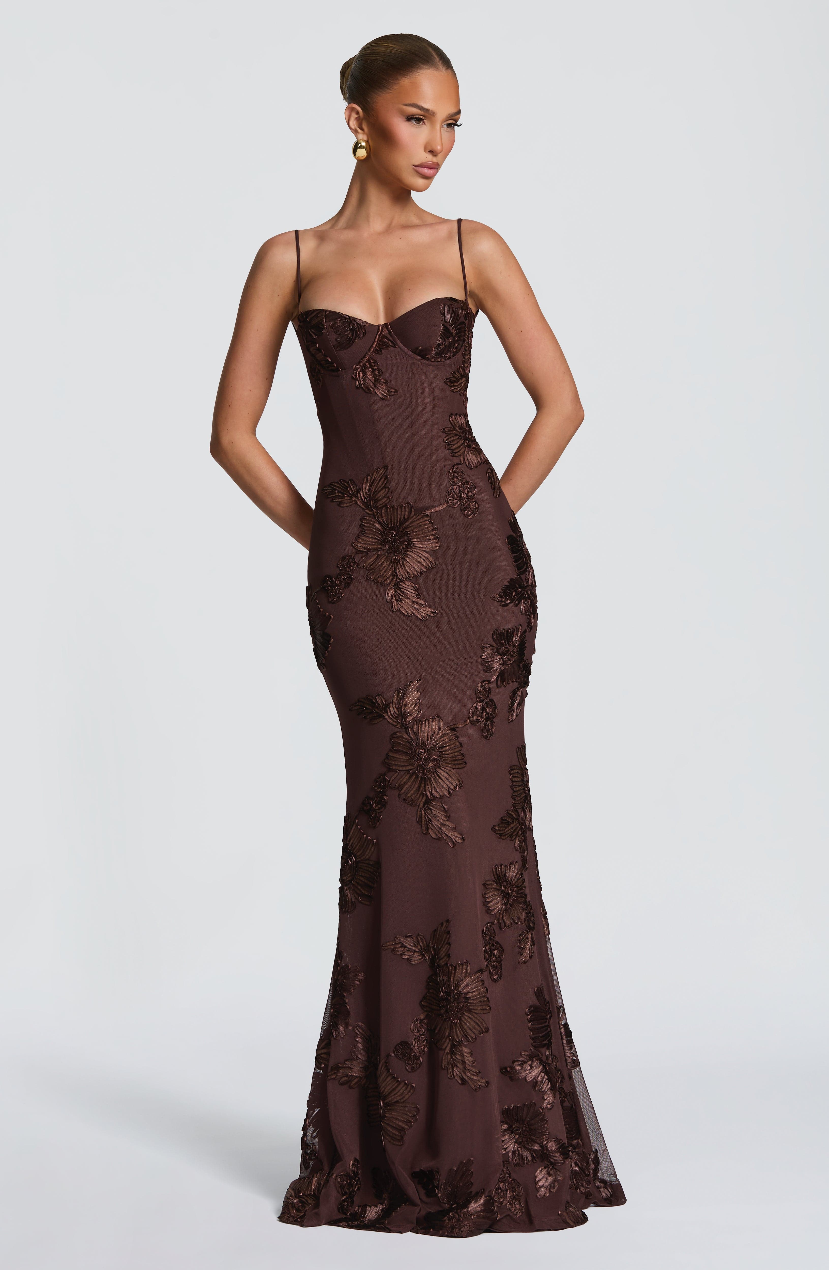 Front view of a woman wearing a dark brown Celestina floral maxi dress with matching dark floral embroidery, featuring a structured bodice and spaghetti straps.