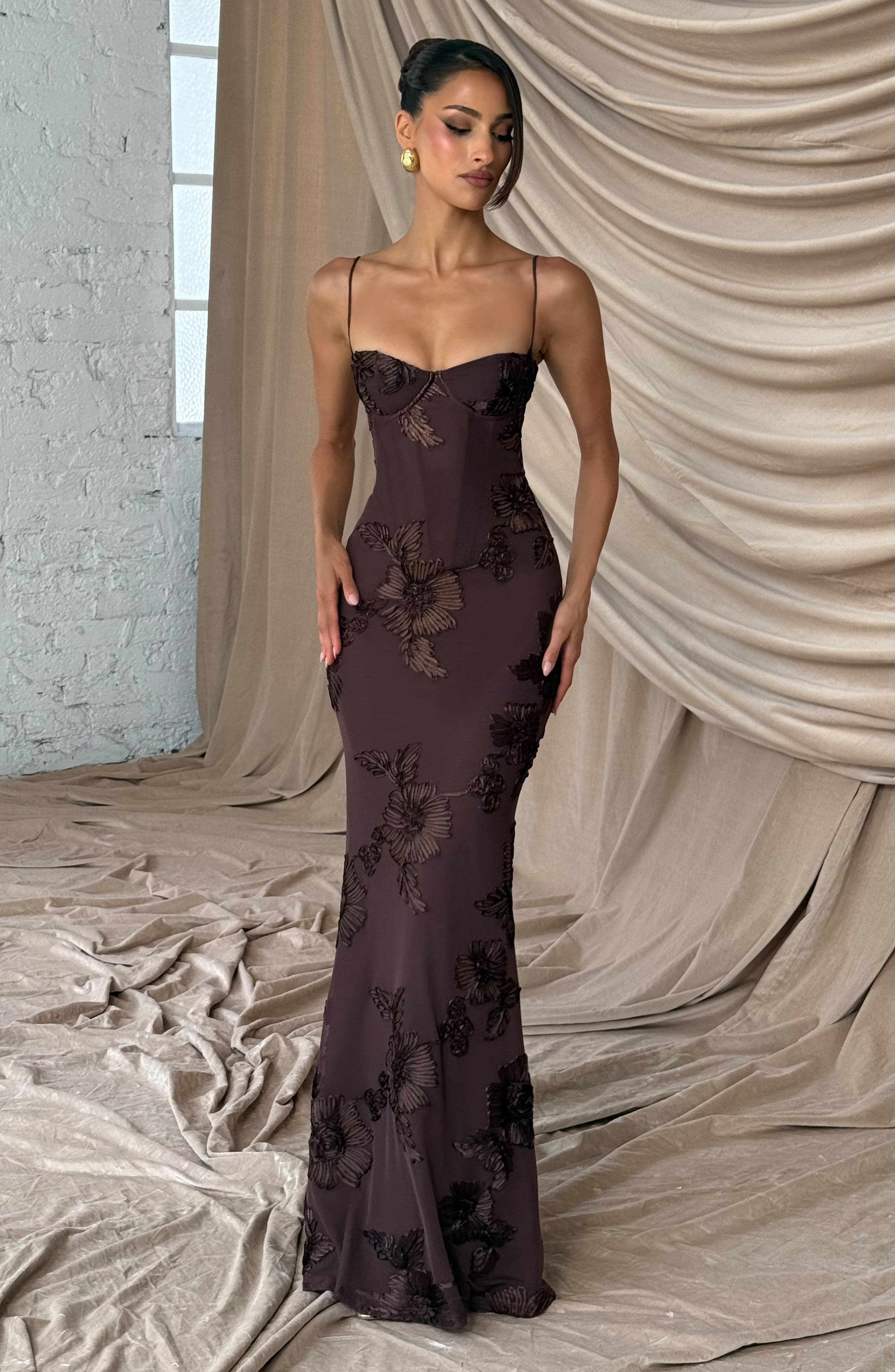 Full body shot of a brown floral maxi dress with subtle floral patterns, standing against a draped fabric backdrop, ideal for plus size formal clothing.