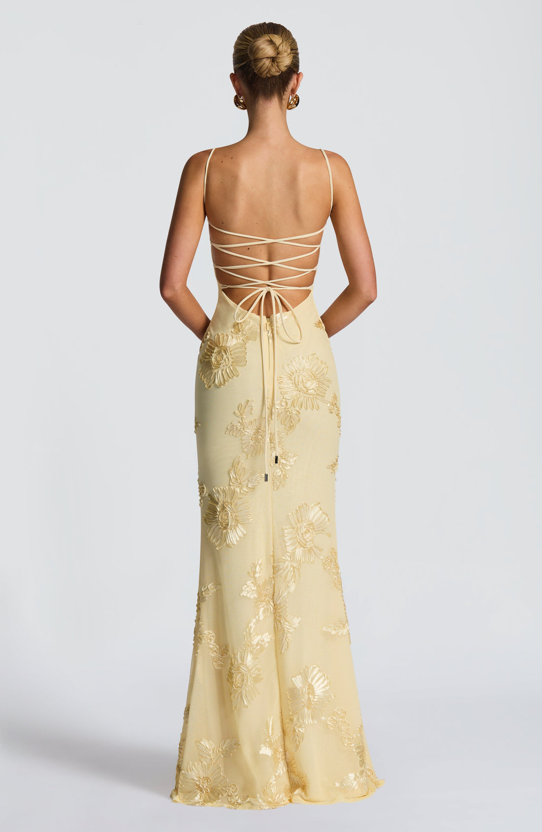 Full-length view of a woman wearing a light yellow Celestina floral maxi dress with delicate spaghetti straps and intricate gold floral embroidery, leaning against a wall.