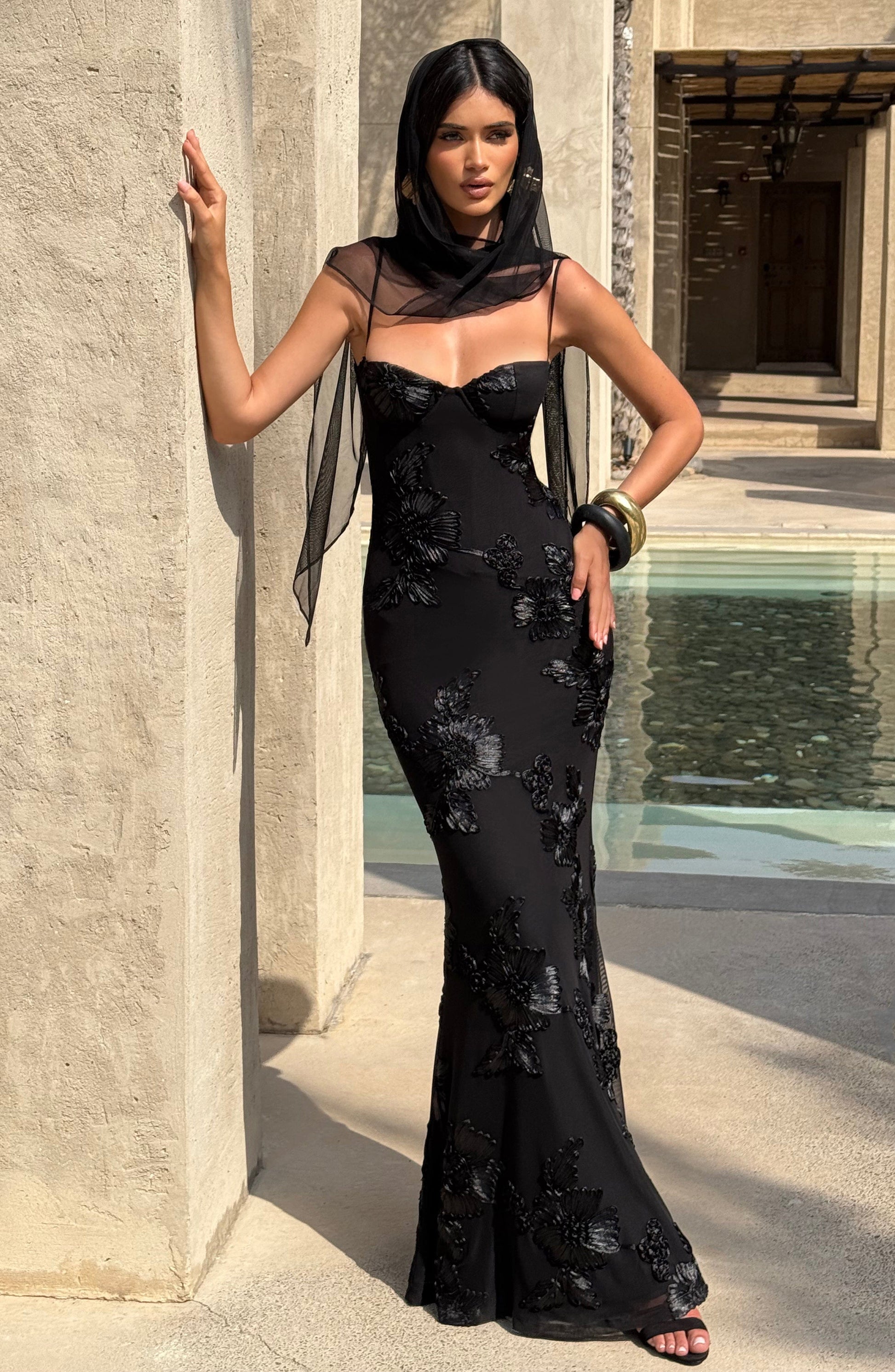 Full length view of a black floral maxi dress with a sweetheart neckline, delicate straps, and intricate black floral embroidery, worn with a sheer black scarf.