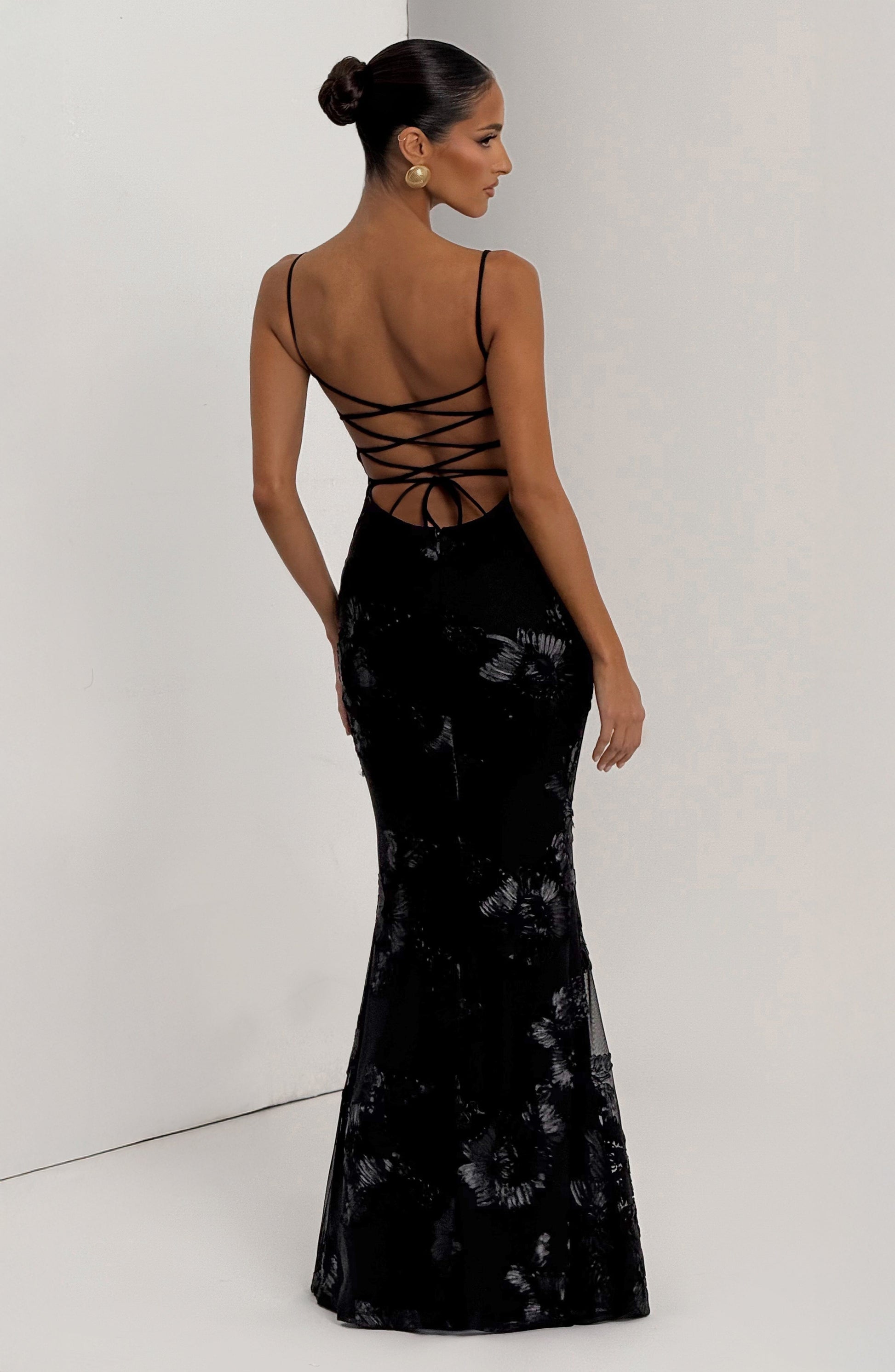 Back view of the black Celestina Floral Maxi Dress, highlighting the lace-up detail and the flowing skirt with scattered floral embellishments.