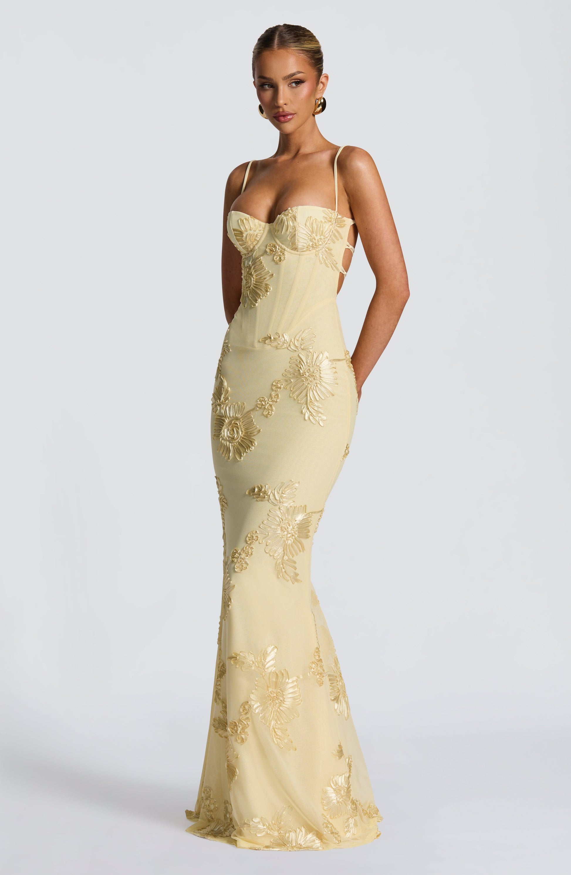 Side profile of a woman wearing the light yellow Celestina floral maxi dress, revealing the lace-up back detail and flowing skirt with gold floral embroidery.