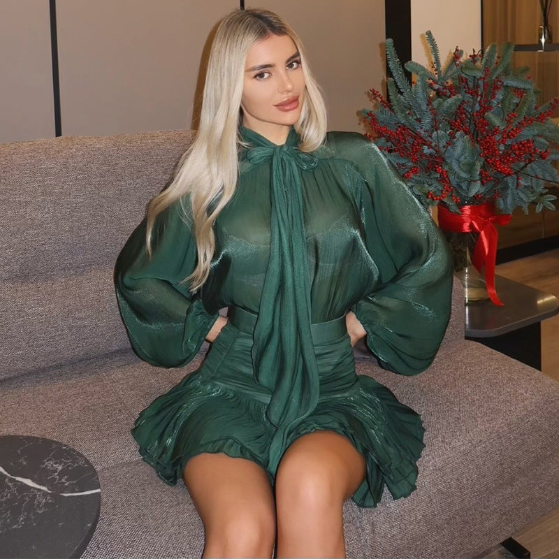 Shimmering dark green Celia Bow Mini Dress with a high neck tie and pleated skirt, long sleeves, on a seated figure.