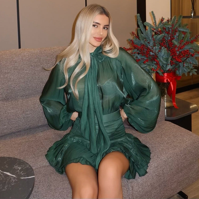 Close-up of the dark green Celia Bow Mini Dress, showcasing the sheer long sleeves and pleated mini skirt on a seated figure.