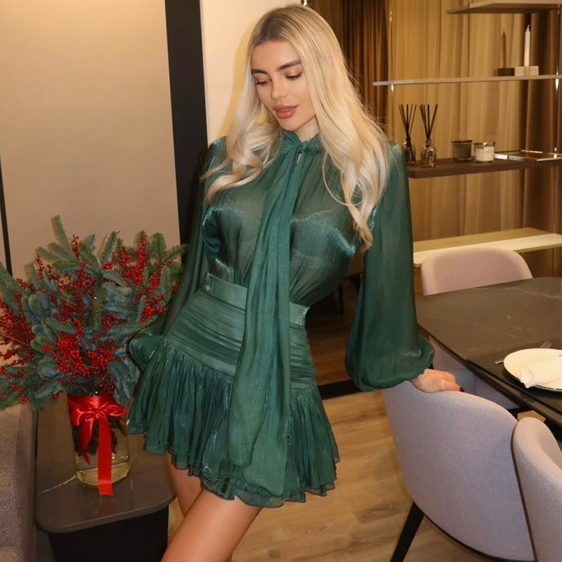Full view of the dark green Celia Bow Mini Dress, featuring a high neck bow and ruffled hem, worn by a standing figure.