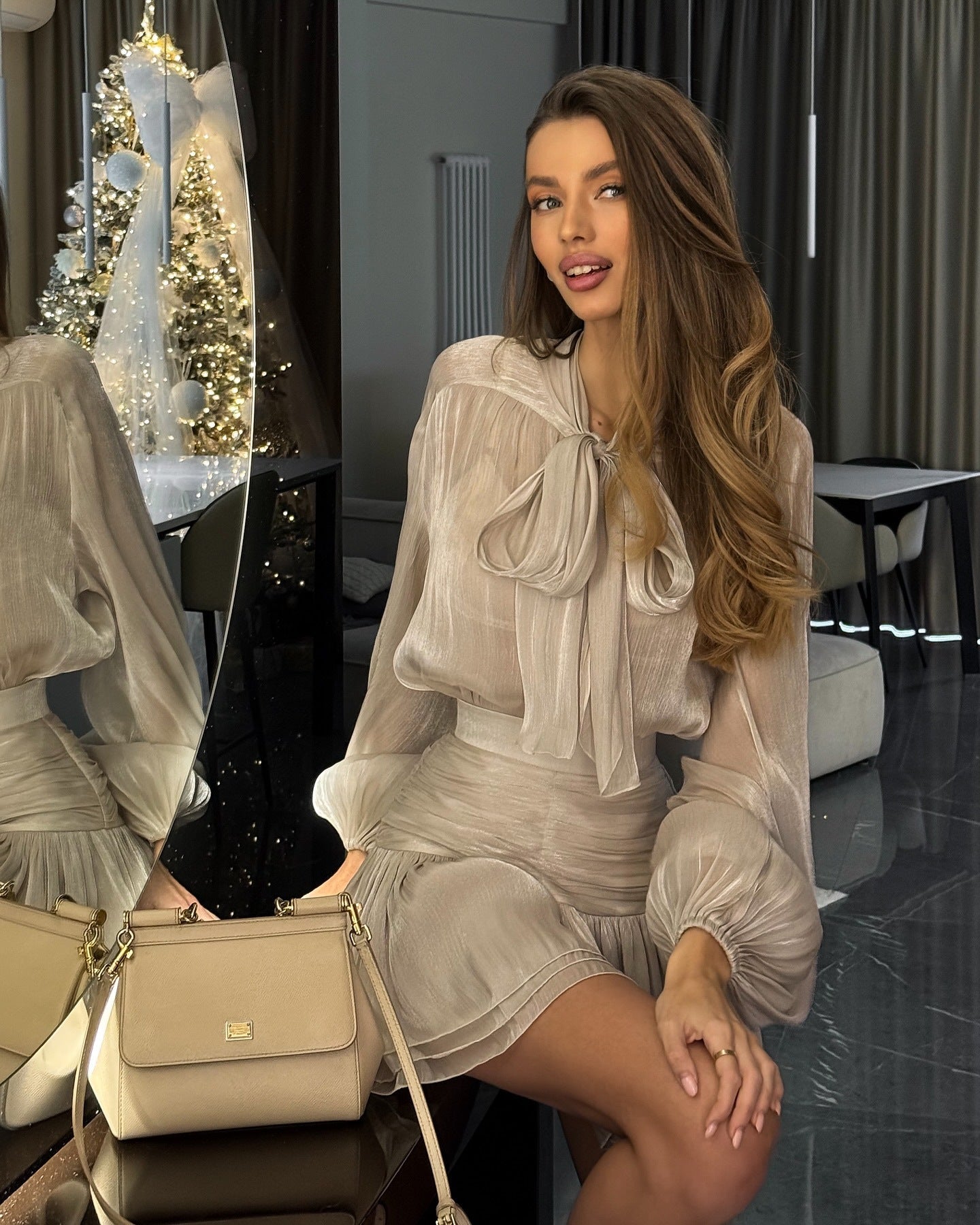 Champagne-colored Celia Bow Mini Dress, showing the sheer fabric and layered pleated skirt, with a handbag beside it.