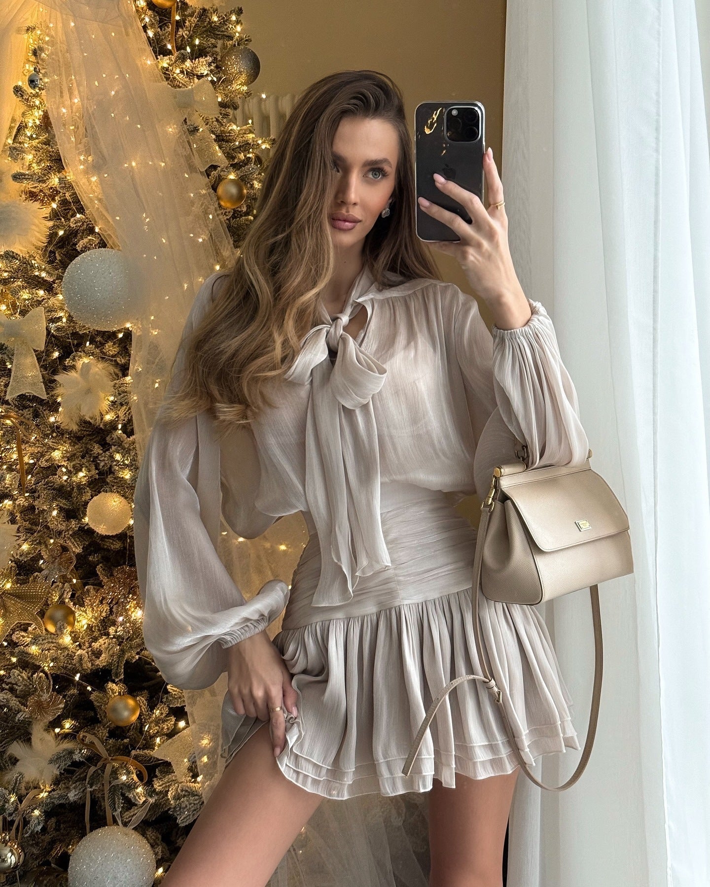 Selfie shot of a woman in a shimmering long-sleeve Celia Bow Mini Dress with a detailed bow neck.