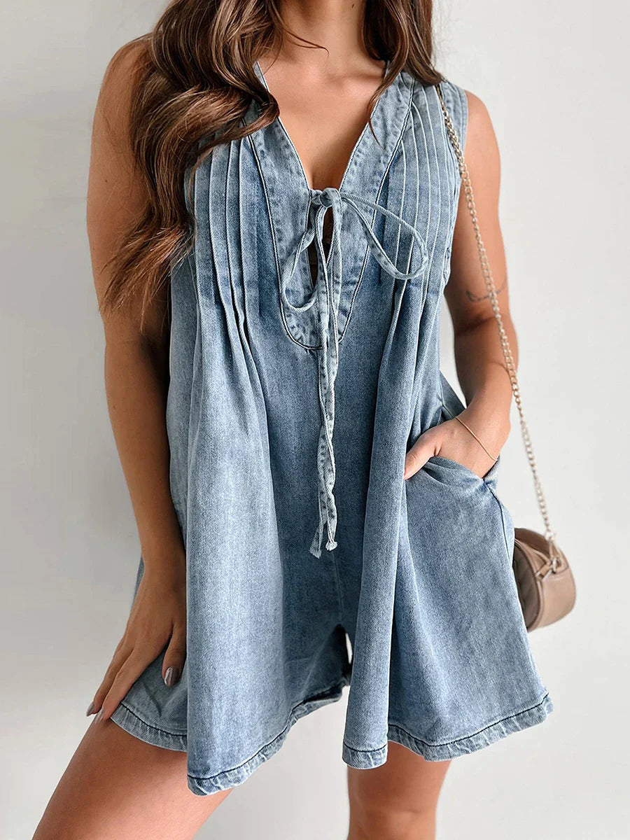 Sleeveless light wash denim mini dress with a v-neck, tie-front closure, pleated details, and side pockets.