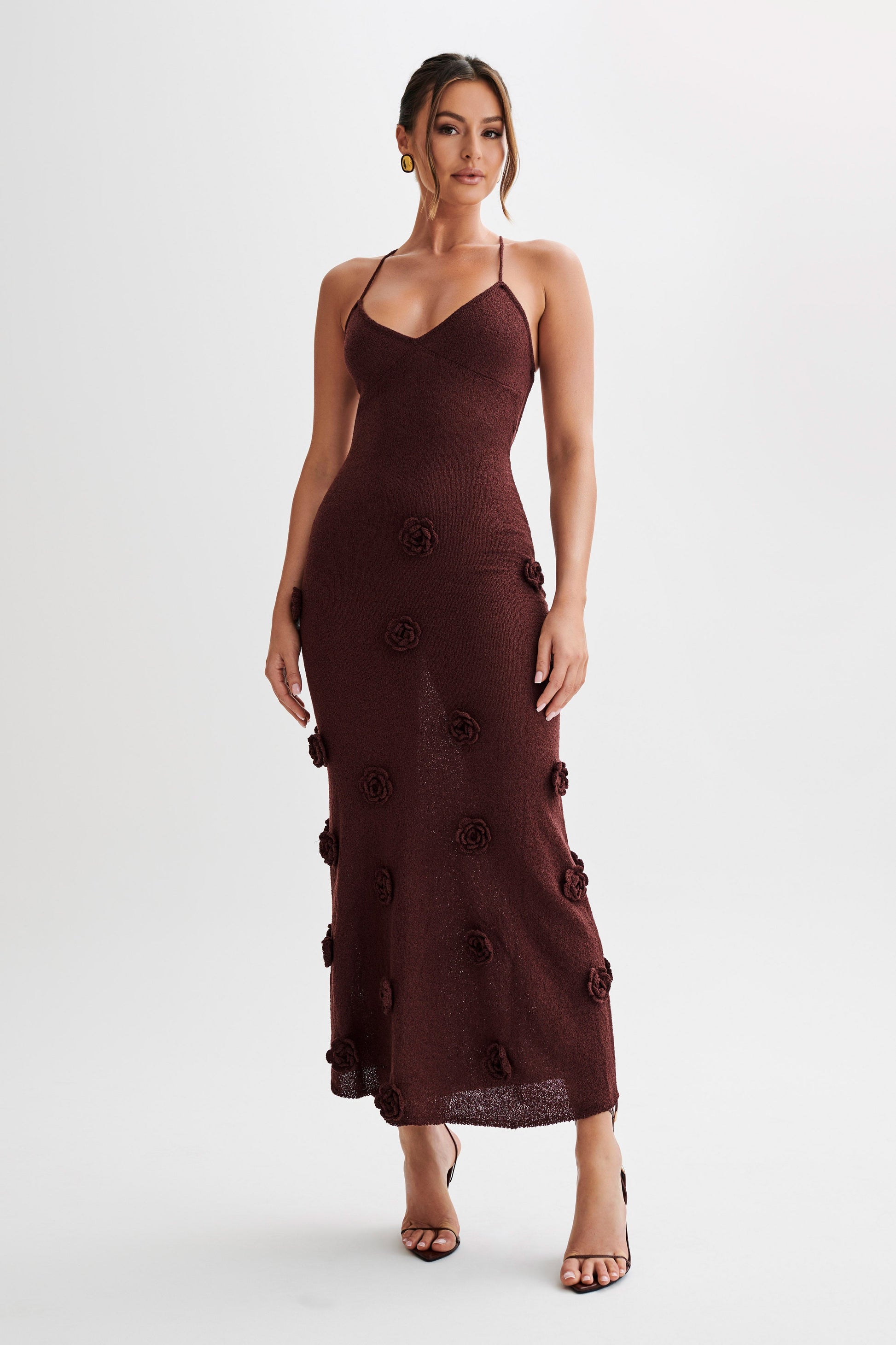 Front view of a dark brown Chelsea Floral Maxi Dress with intricate rose appliques, styled with open-toe heels.