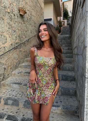 Multicolored mosaic sequin mini dress with square neckline and side slit for women, worn by a model smiling on stone steps.