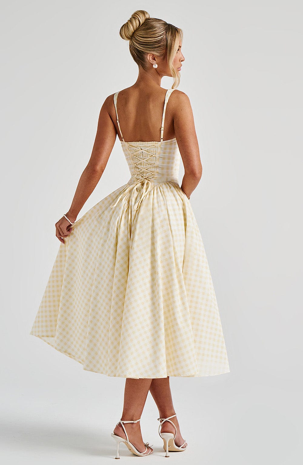 Rear view of a light yellow gingham midi dress, showcasing its spaghetti straps, lace-up back, and full skirt, with a hand gently lifting the hem.