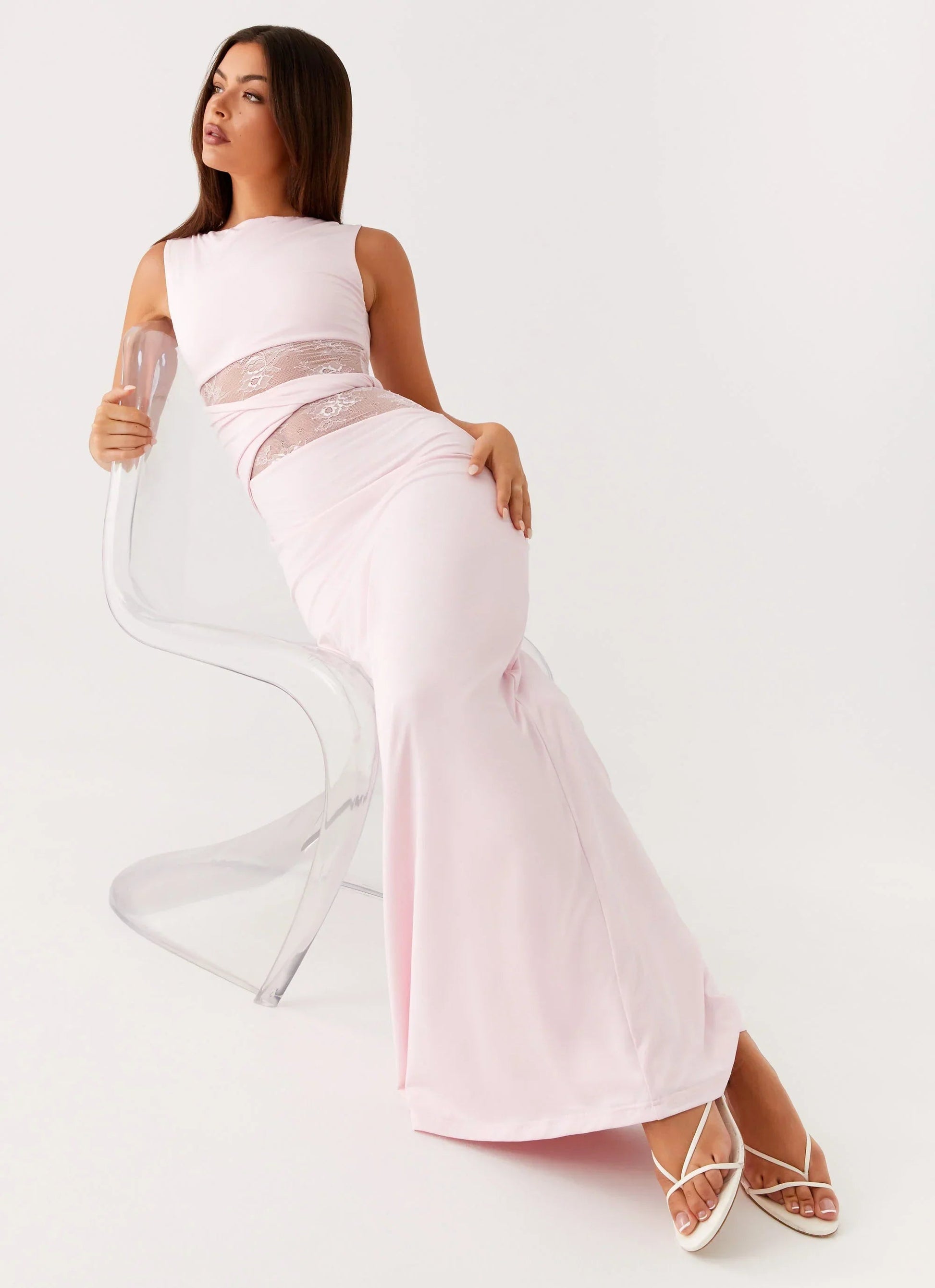 Light pink Clarisse lace maxi dress with sheer waist details, worn with white heels, seated