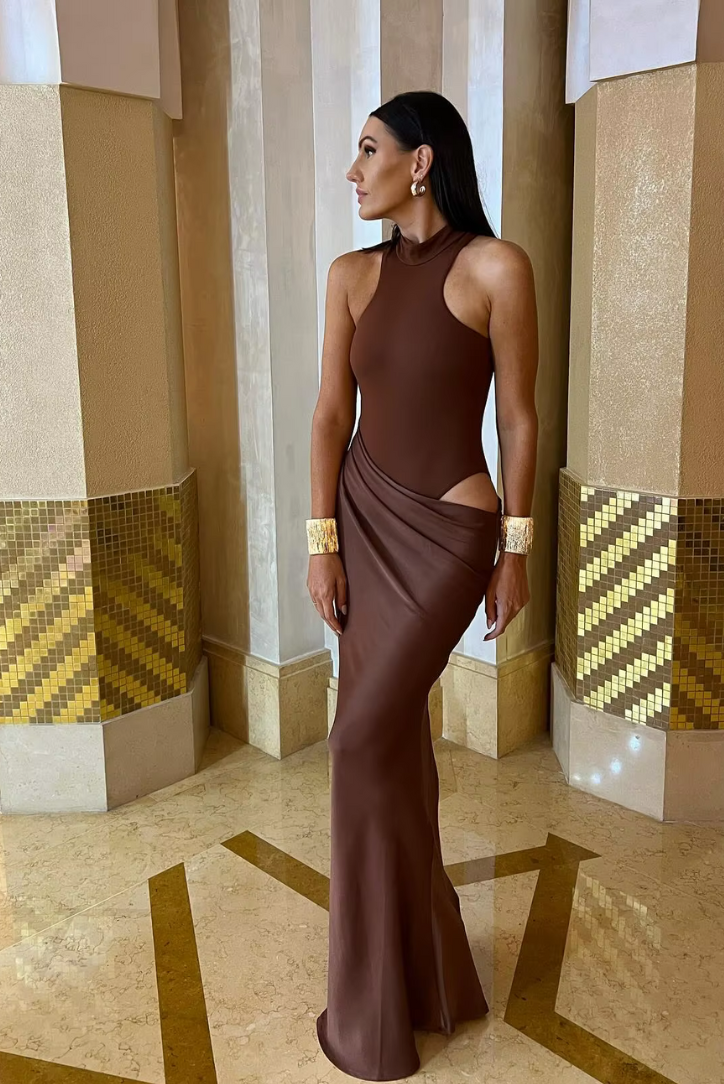 Elegant brown cutout maxi dress with a draped skirt, worn with gold cuff bracelets and earrings.
