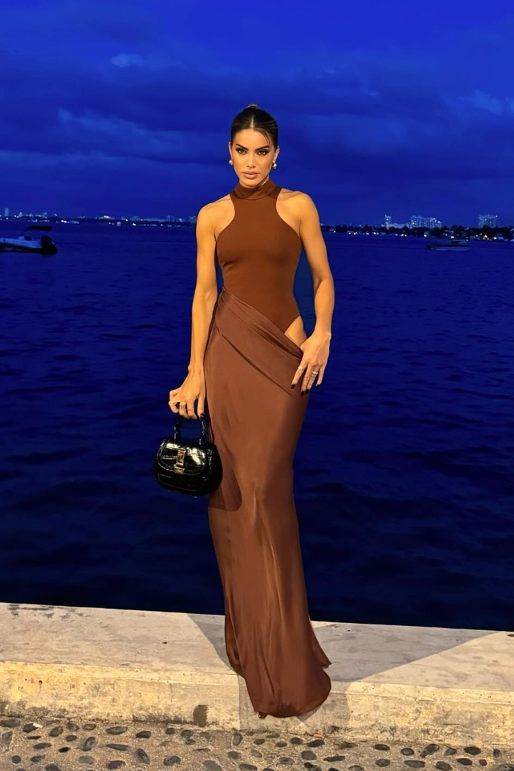 Brown cutout maxi dress with a high neck, styled with a black handbag, against a dark blue water background.