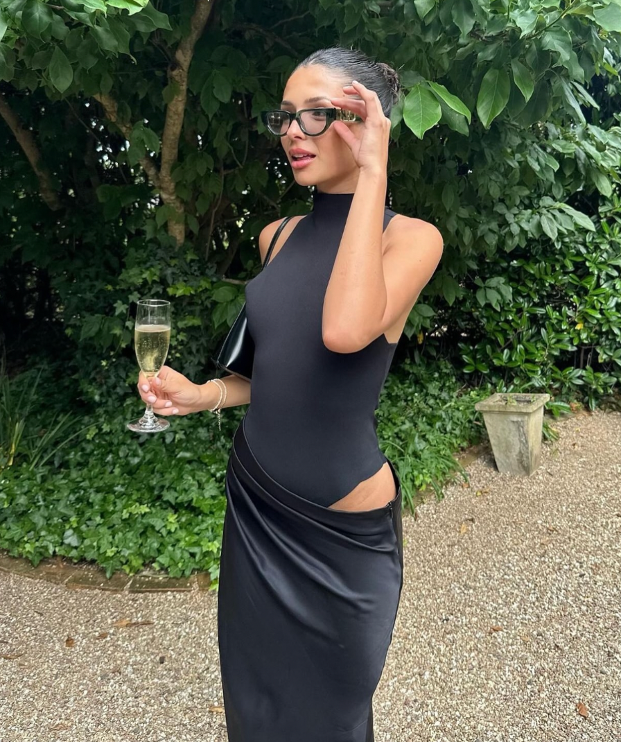 Black cutout maxi dress with a high neck, holding a champagne glass, wearing dark sunglasses.